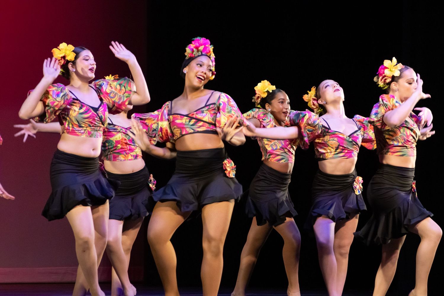 A group of female dancers in tropical floral tops and black skirts perform a lively Latin dance.
