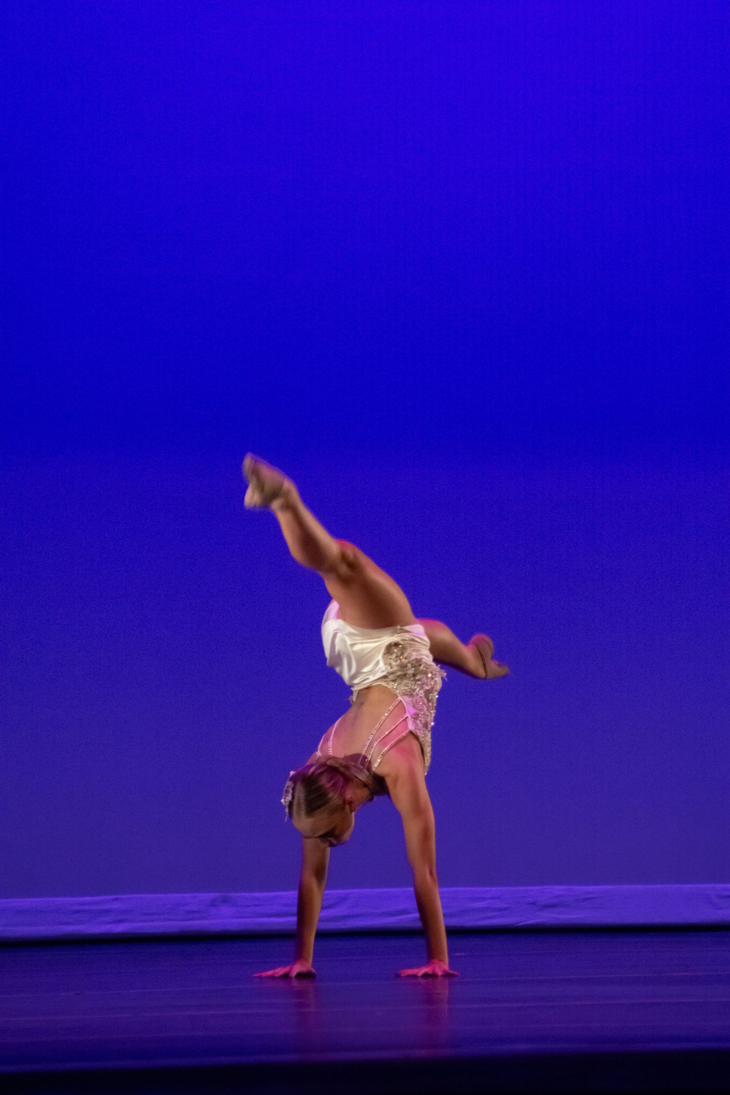 A young dancer performs a one-handed handstand on a blue-lit stage wearing a white costume.