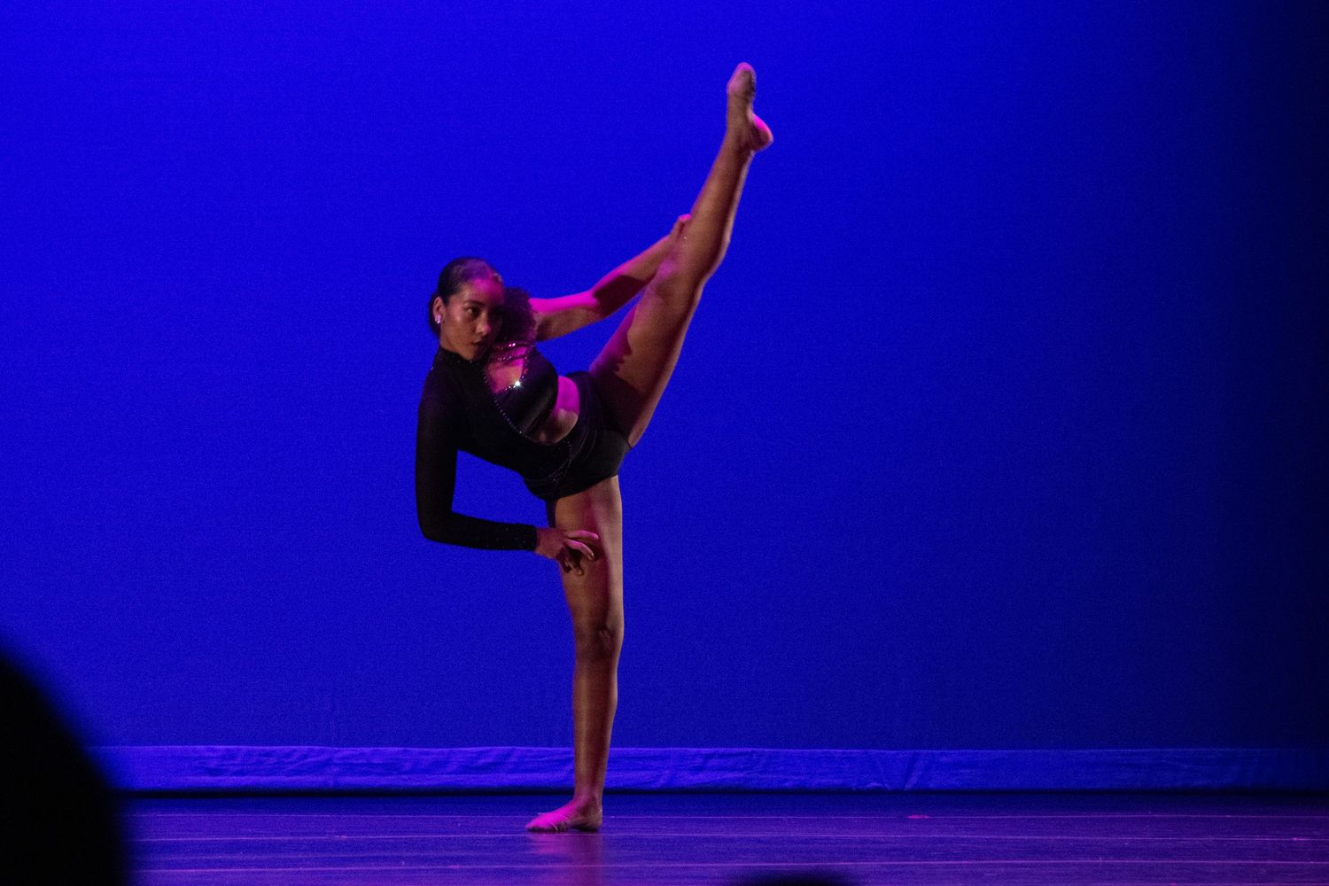 A skilled dancer performs a high leg extension on a blue-lit stage in a black costume.