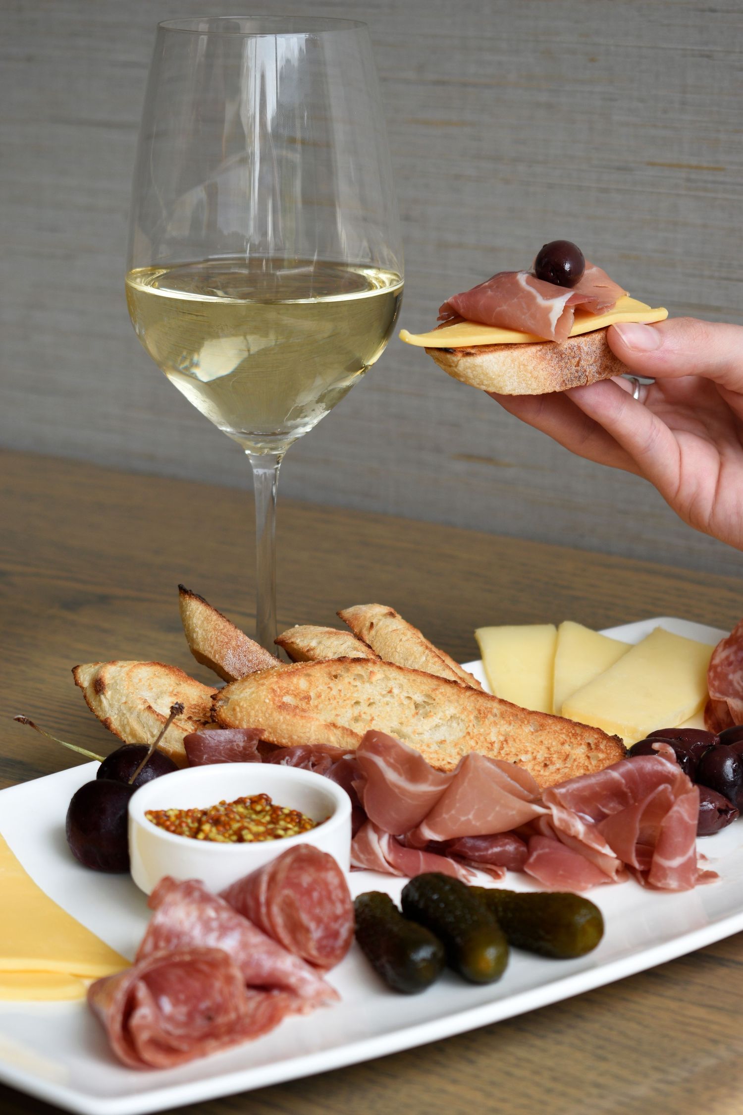 Hand holding bruschetta with cheese over a charcuterie board of meats, olives, and pickles with white wine.