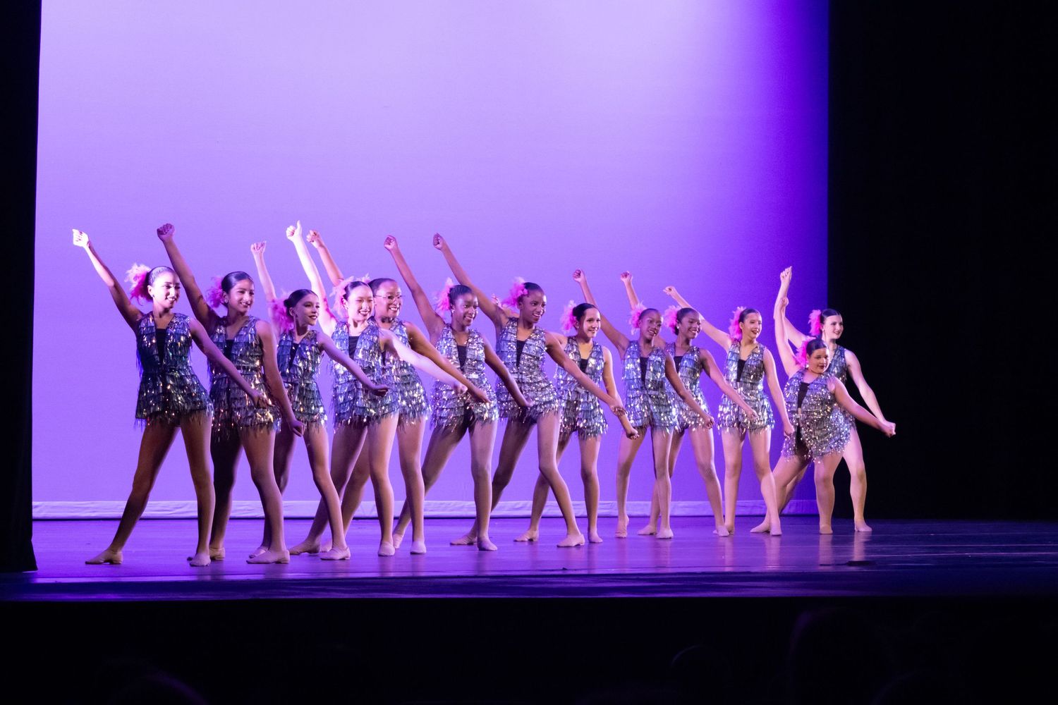 A line of dancers in sparkly costumes perform on a purple-lit stage.