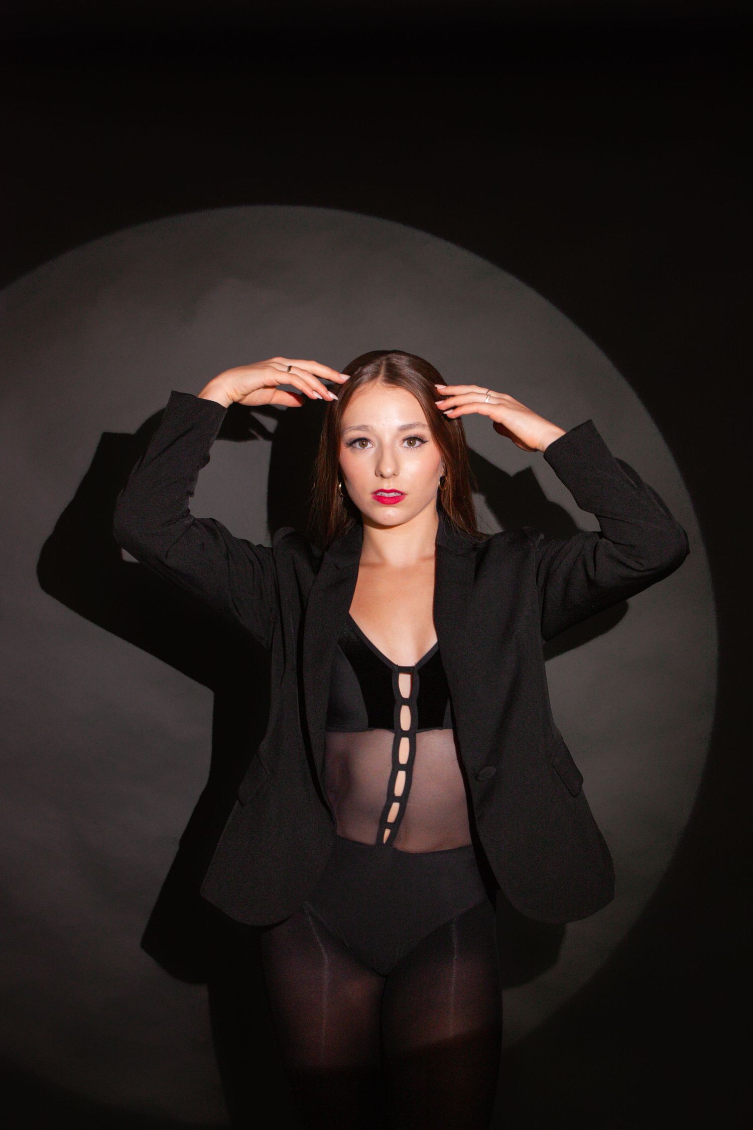 Woman in black blazer and sheer bodysuit posing dramatically against dark background with spotlight circle.