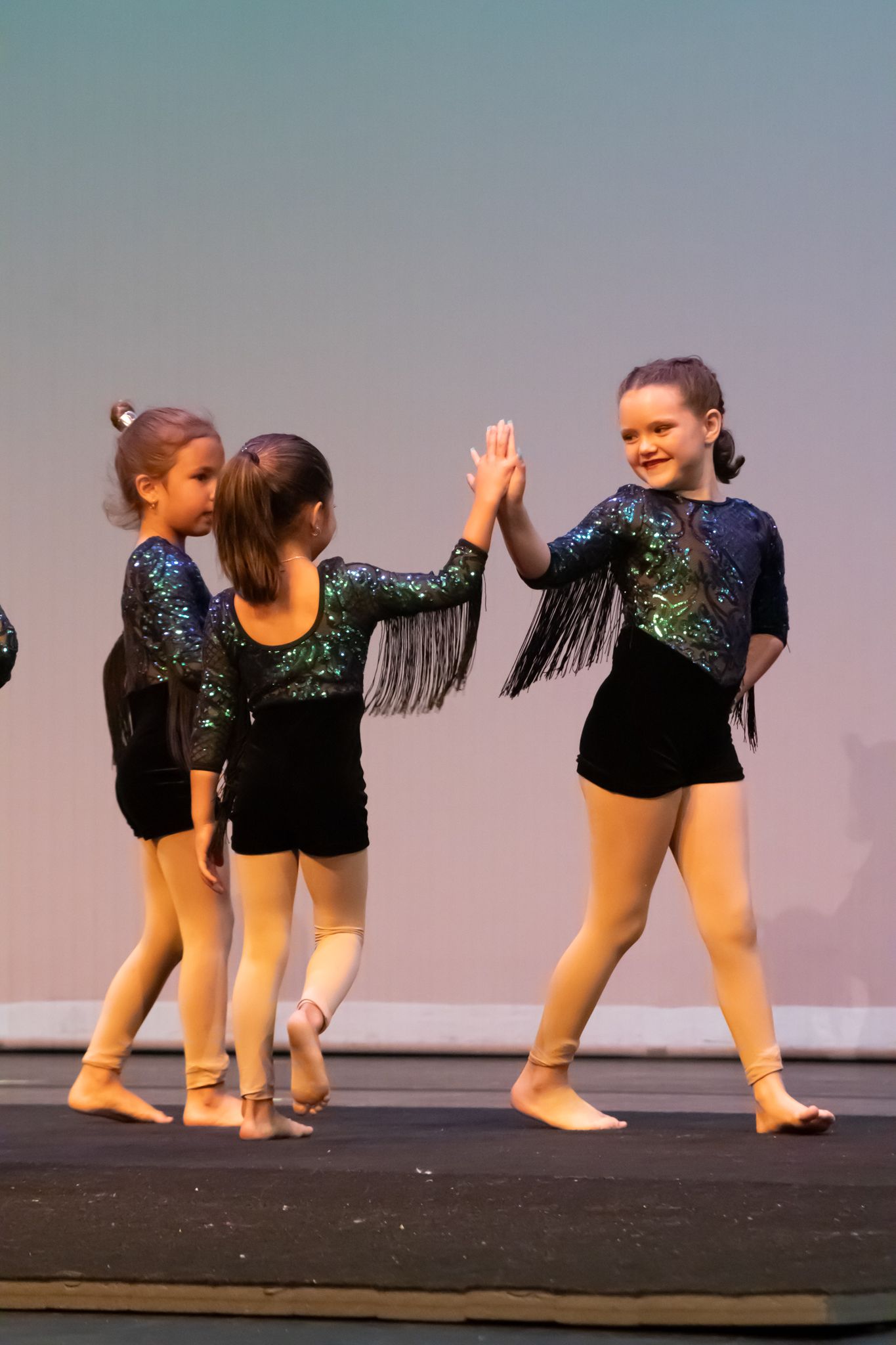 Three young girls in sequined dance costumes high-five on stage during a performance.