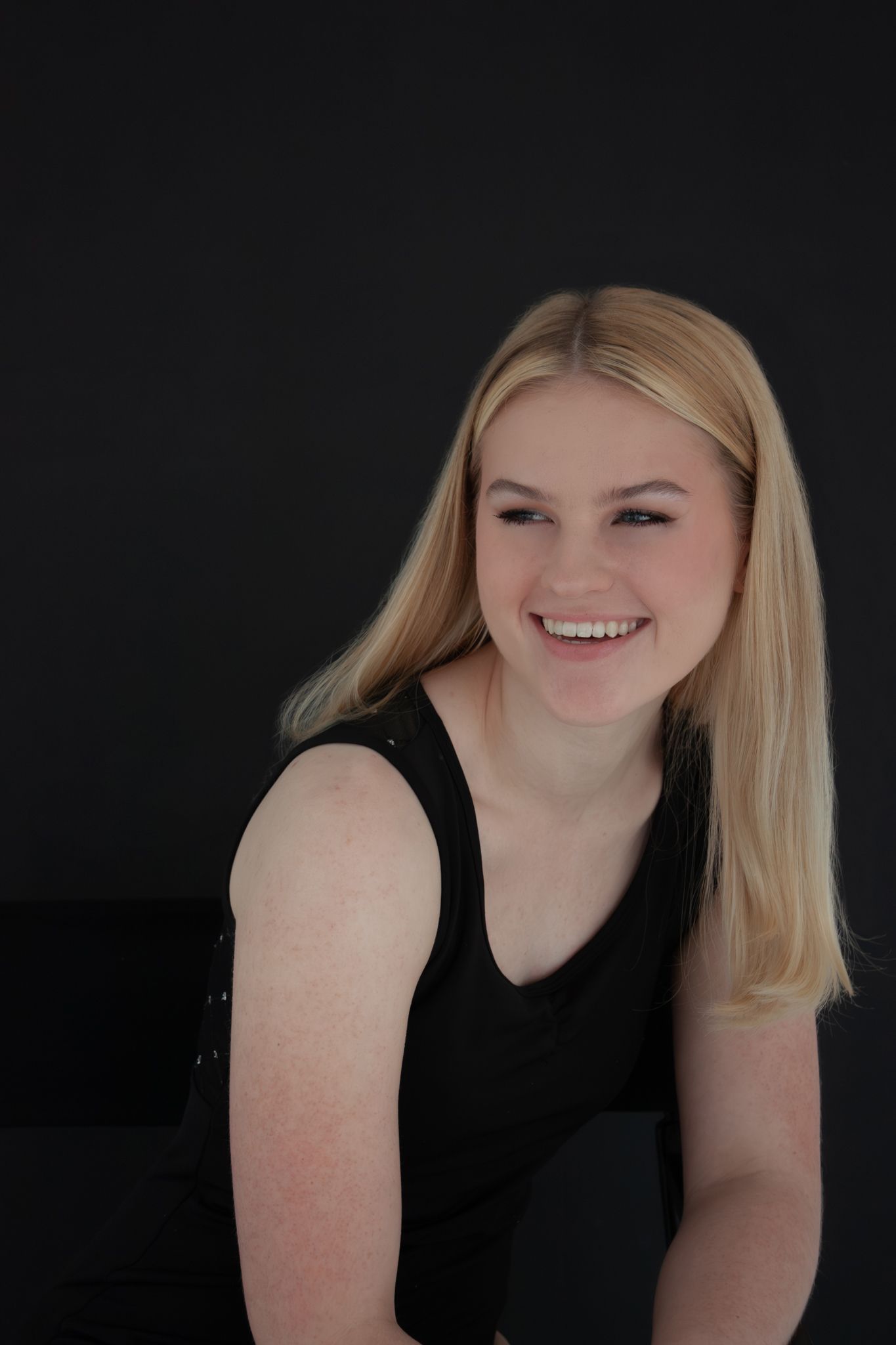 Smiling blonde young woman in black sleeveless top laughs against a dark background.