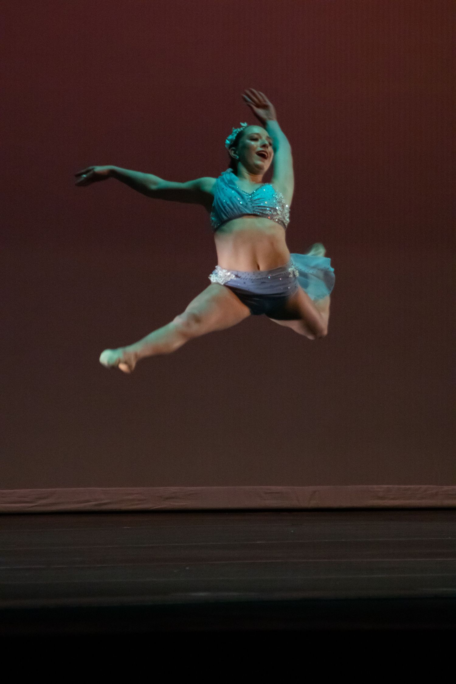 A smiling dancer in a teal and white costume performs a joyful leap on a red-lit performance stage.