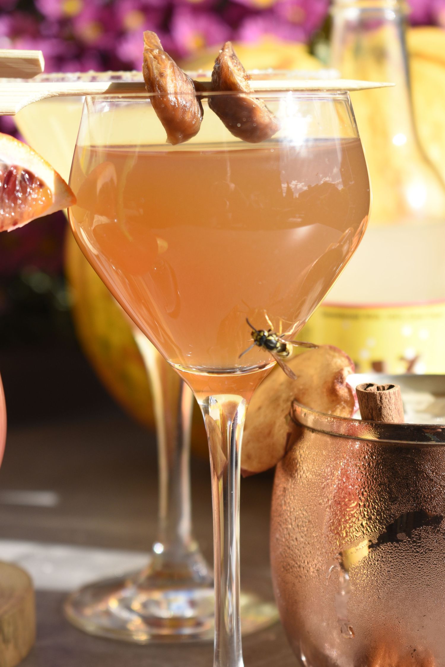 Pink grapefruit cocktail garnished with dried fruit skewer in a stemmed glass with copper mug nearby.