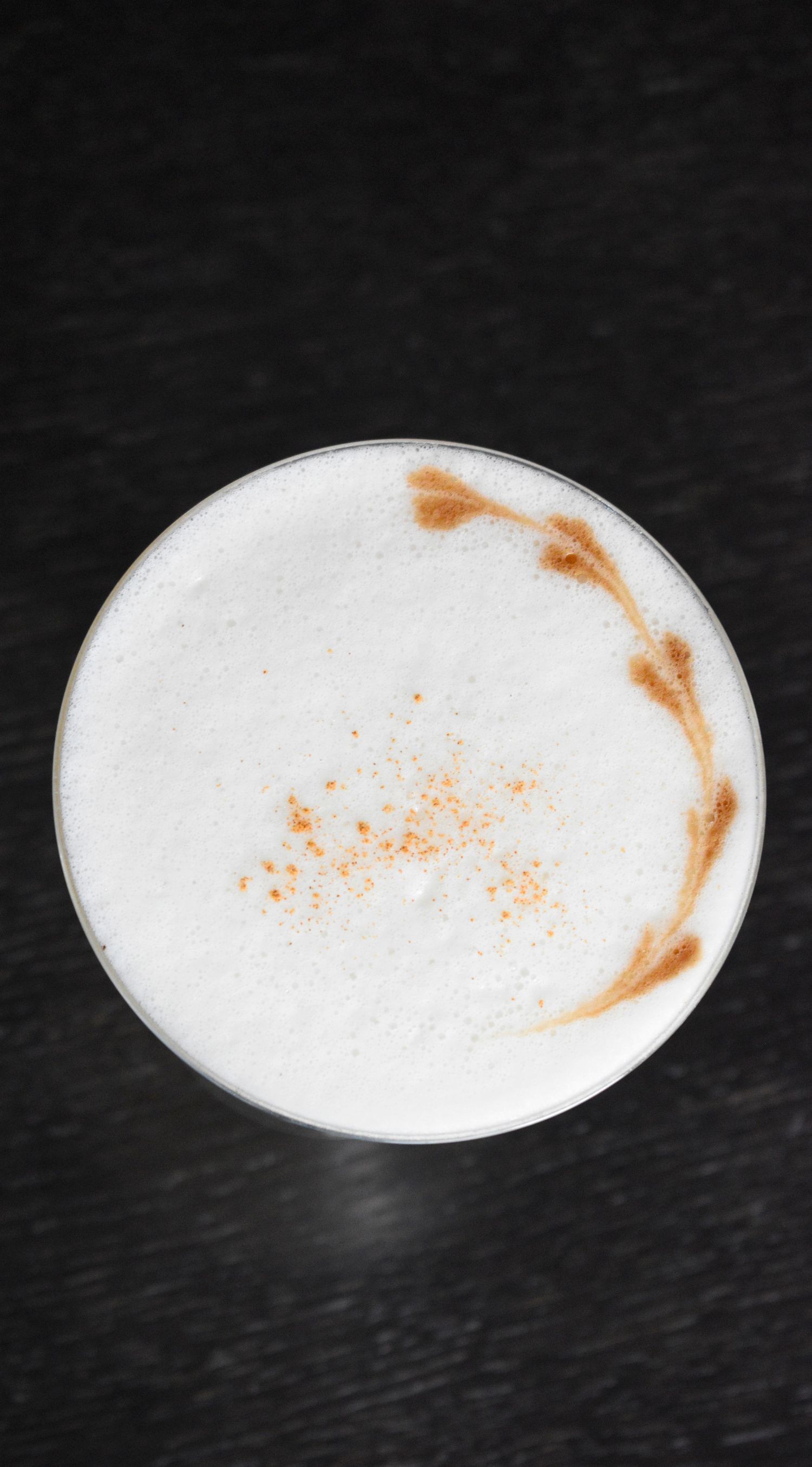 An overhead view of a frothy white cocktail dusted with cinnamon spice in a coupe glass.