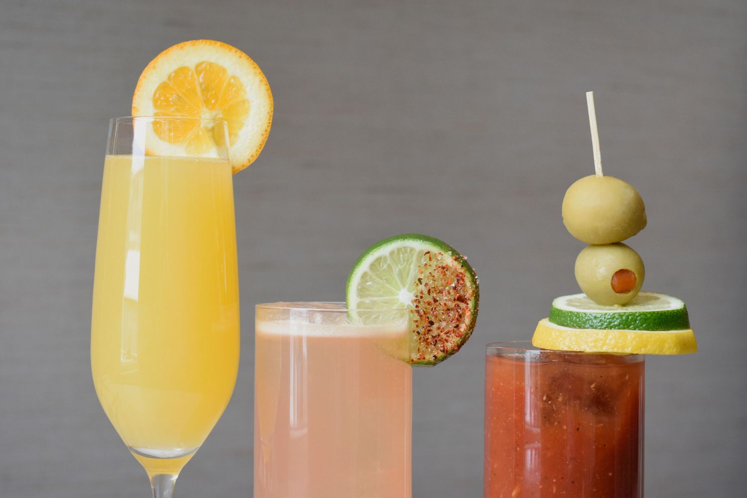 Three brunch cocktails including a mimosa, paloma, and Bloody Mary with citrus and olive garnishes.