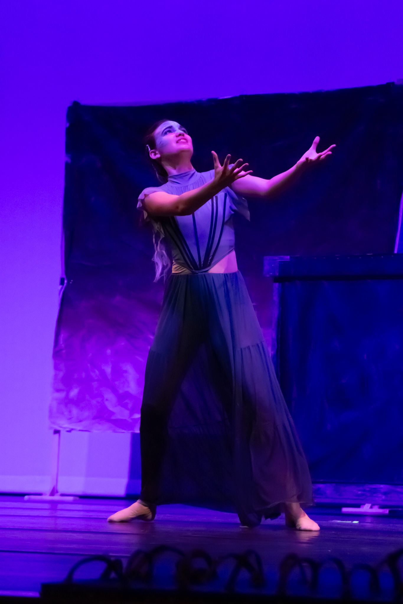 A solo dancer in a flowing grey costume performs an expressive contemporary routine on a purple-lit stage.