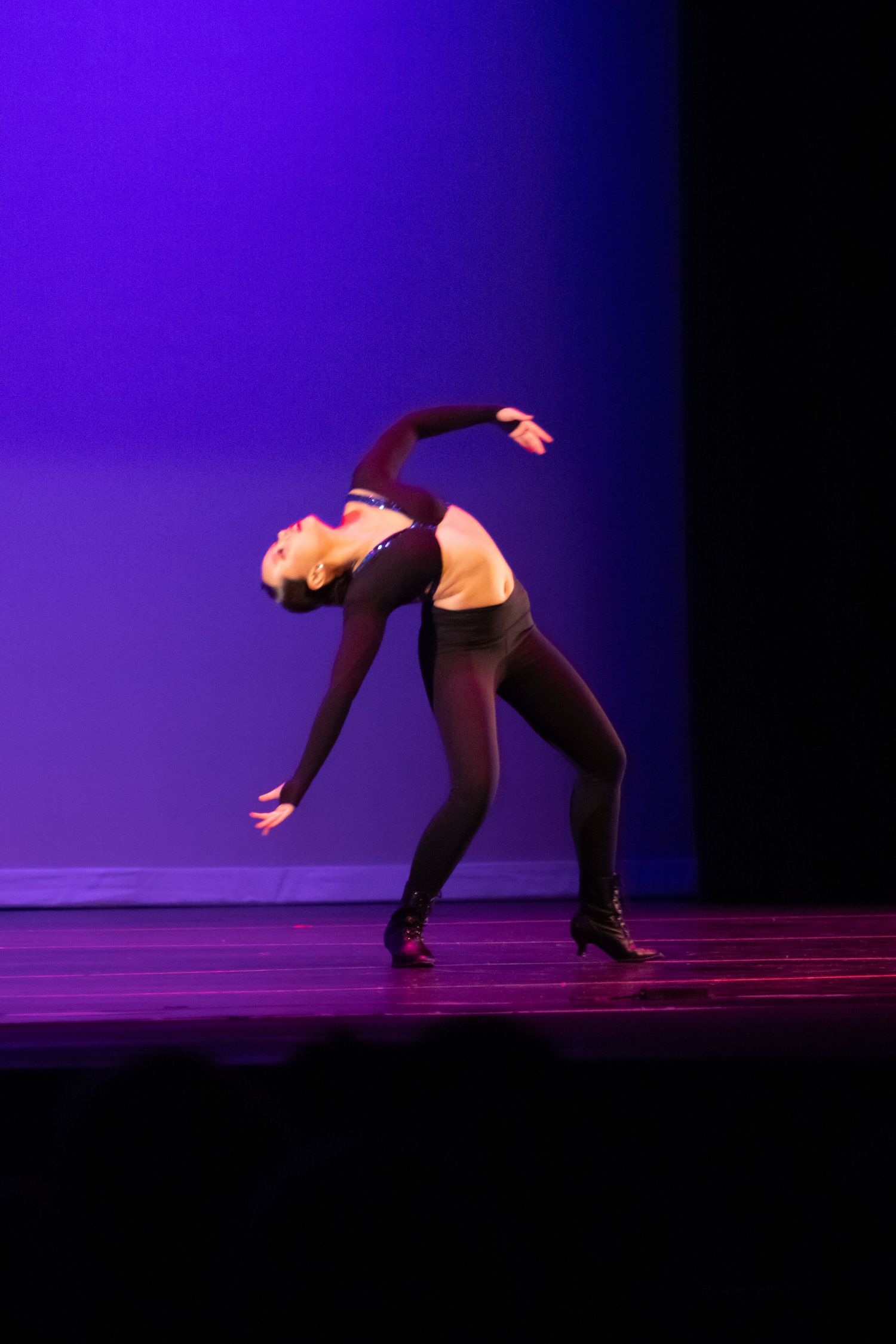 A dancer in black attire performs a dramatic back arch on stage under purple and blue lighting.