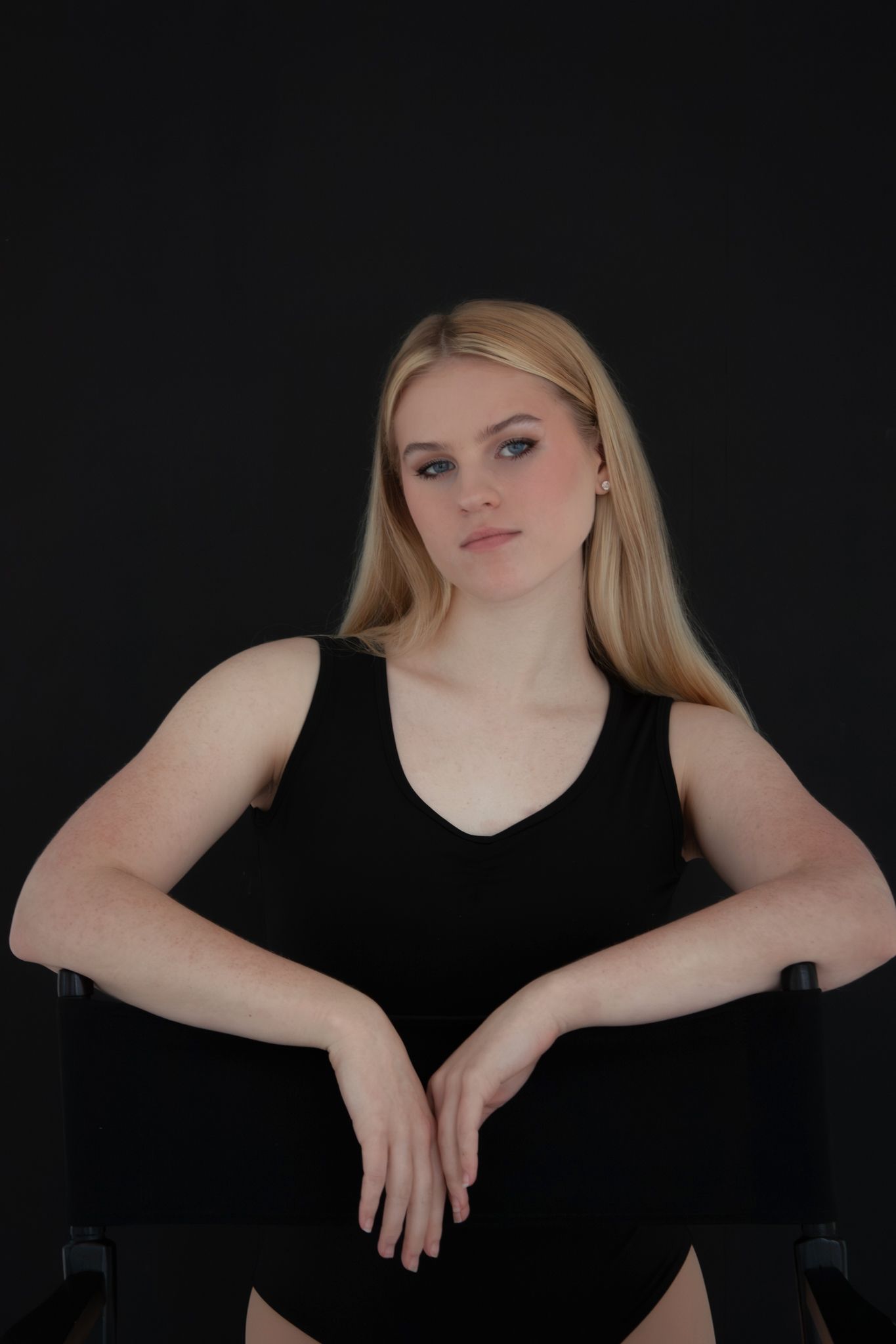 Young blonde woman in black sleeveless top sitting calmly with hands folded on dark background.