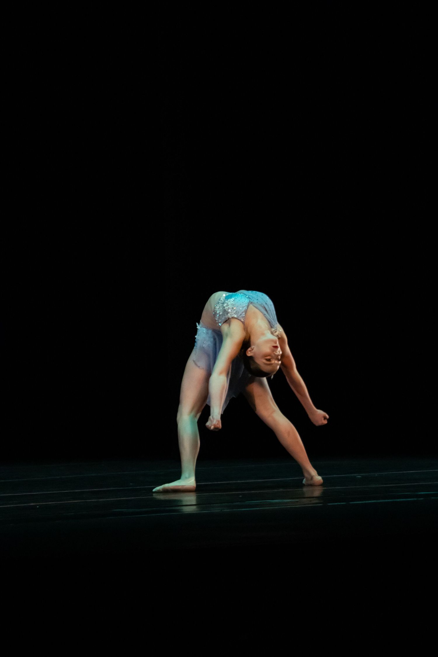 A dancer in a blue costume performs a deep backbend with head lowered on a darkened stage.