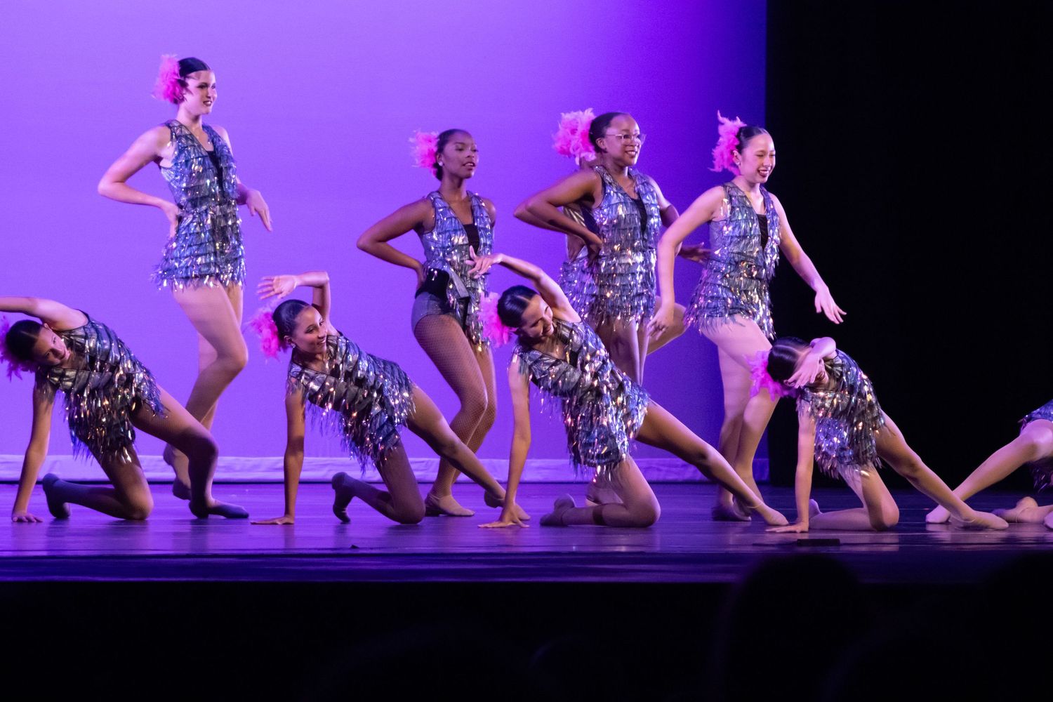 Group of dancers in silver sequin costumes perform an energetic routine on a purple-lit stage.