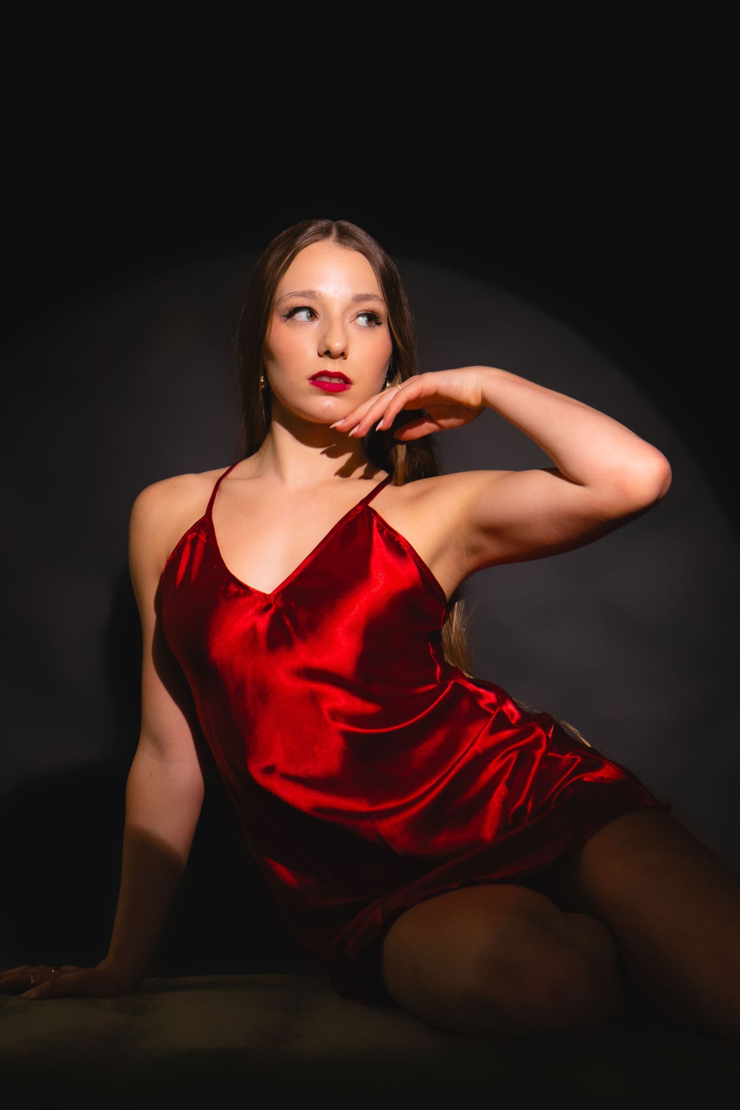 Elegant woman in red satin slip dress posing gracefully against a dark dramatic studio background.