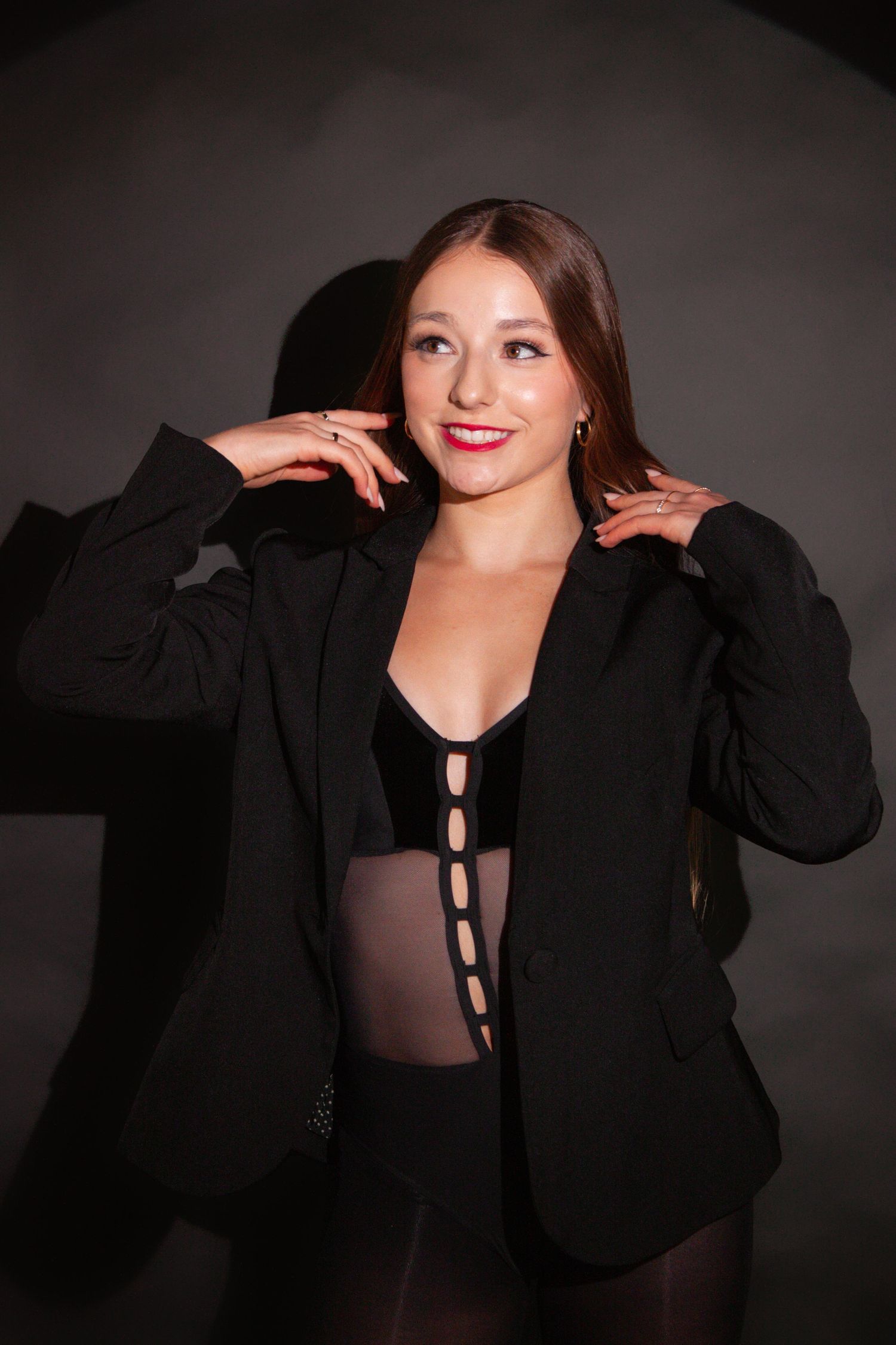Young woman in black blazer and bodysuit smiles confidently against a dark gray studio background.