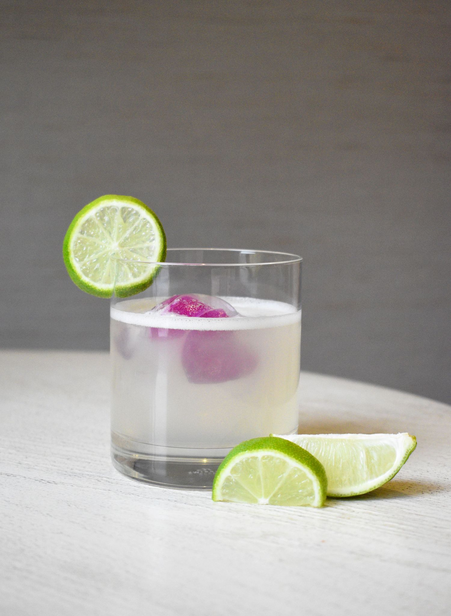 Milky white cocktail in a rocks glass with a pink ice sphere and fresh lime wheel and wedge garnishes.