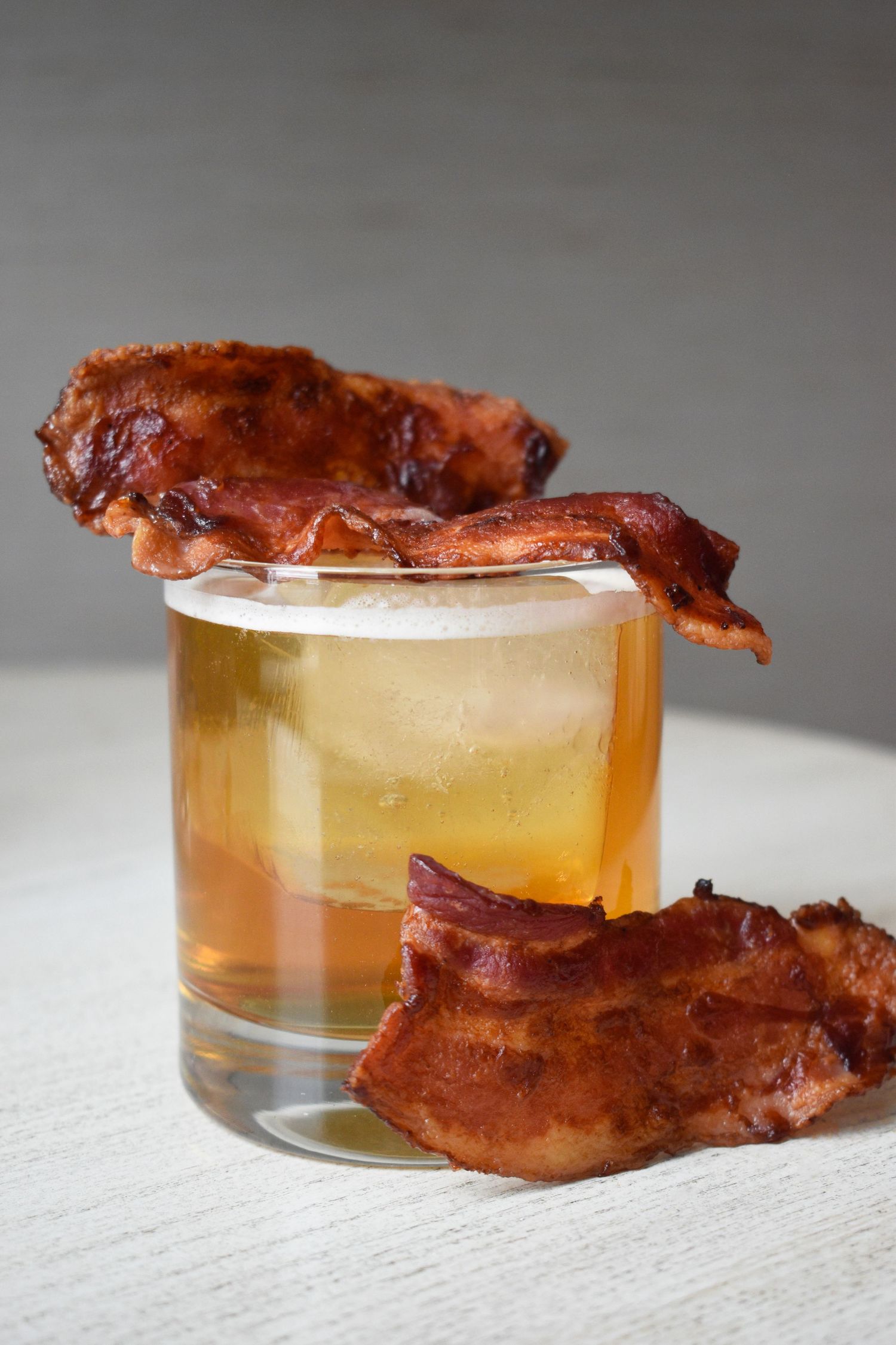 Rustic whiskey cocktail on the rocks garnished with two strips of crispy candied bacon on a white surface.