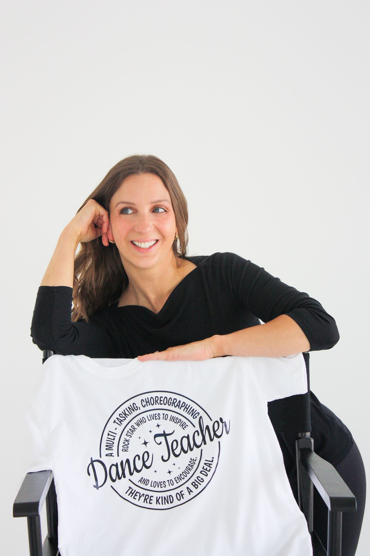 Smiling woman holding white Dance Teacher tshirt, seated in a director's chair against white background. Branding photography in Tampa, FL.