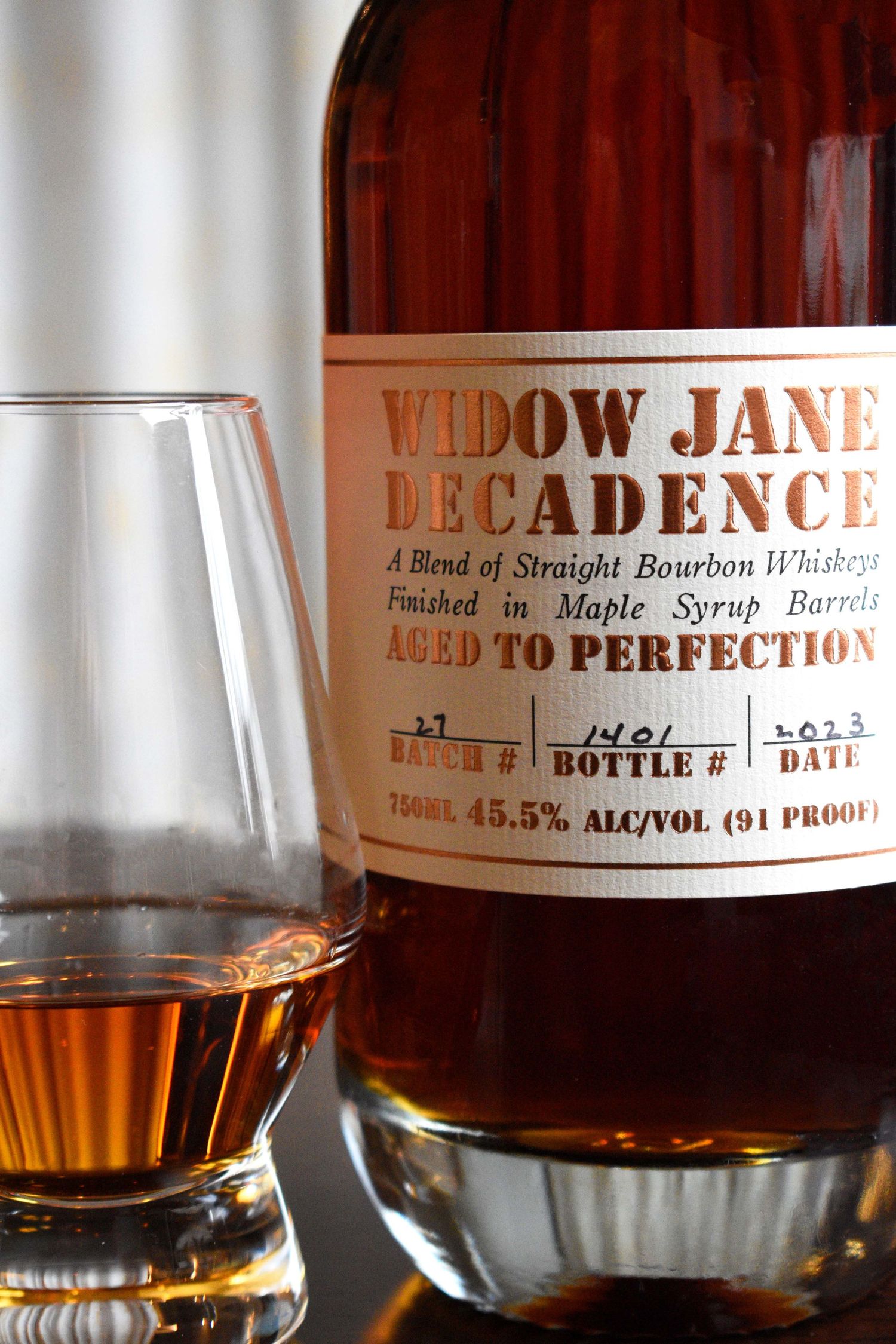 Widow Jane Decadence bourbon whiskey bottle aged in maple syrup barrels beside a Glencairn glass of amber spirit.