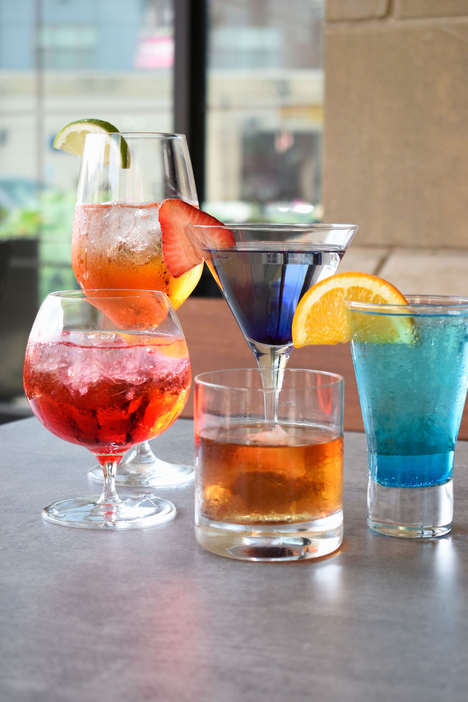 Five colorful cocktails with citrus and fruit garnishes displayed on a bar table.
