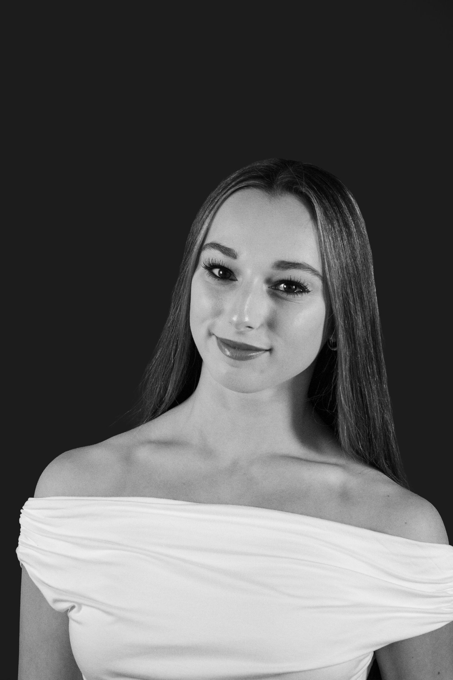 Black and white portrait of a young woman in an off-shoulder white dress, smiling softly against a dark background.