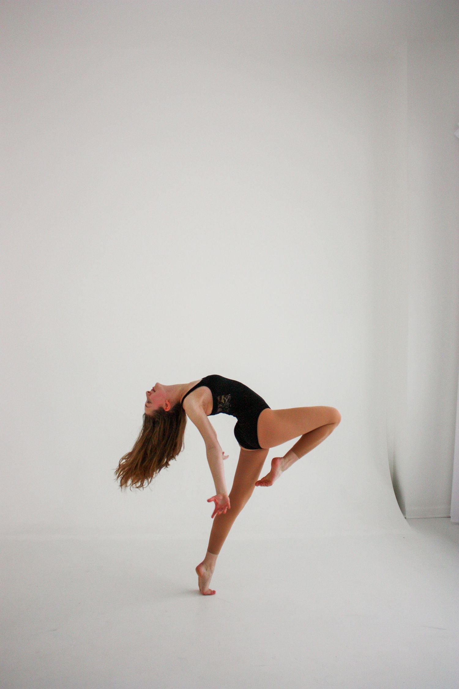 A flexible dancer in a black leotard performs a dramatic backbend on one leg against a white studio background.