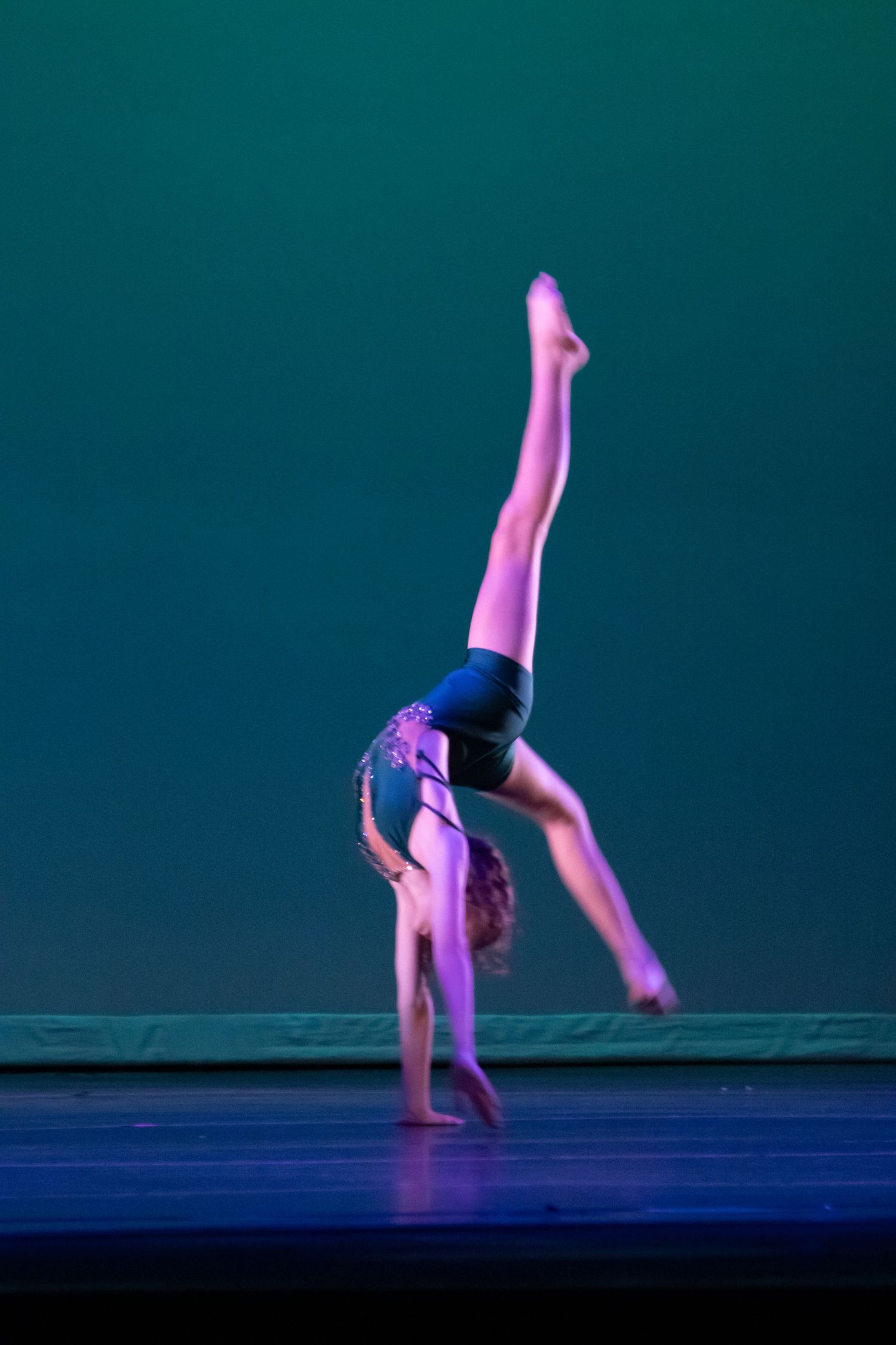 A young dancer executes a one-handed cartwheel with legs split high against a teal stage backdrop.