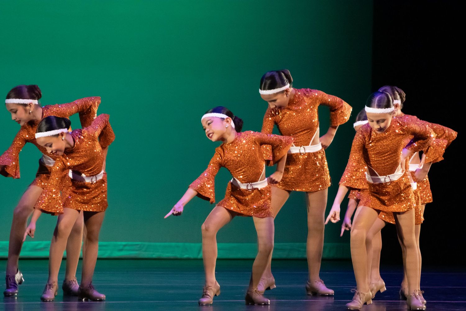 Young dancers in orange sequined costumes perform a synchronized routine on a green-lit stage.