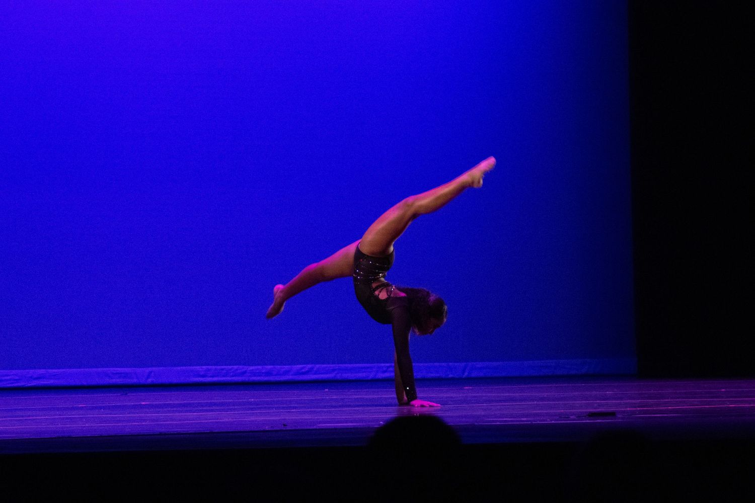 A gymnast performs a one-handed handstand on a dimly lit stage with a blue backdrop.