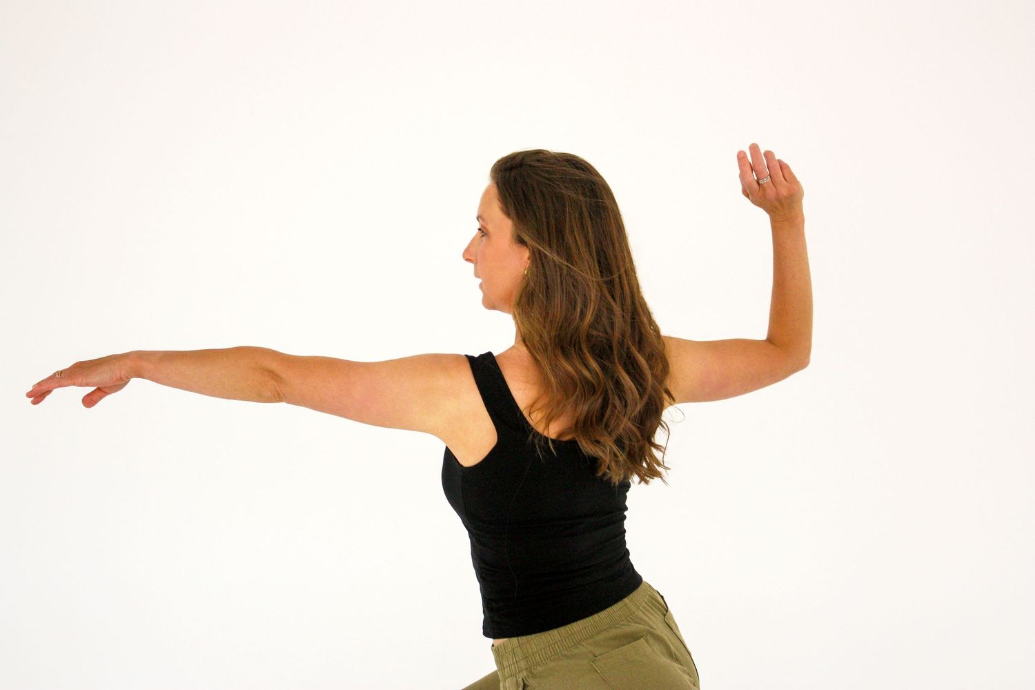 Woman in black tank top and olive pants dancing with arms extended in a white studio.