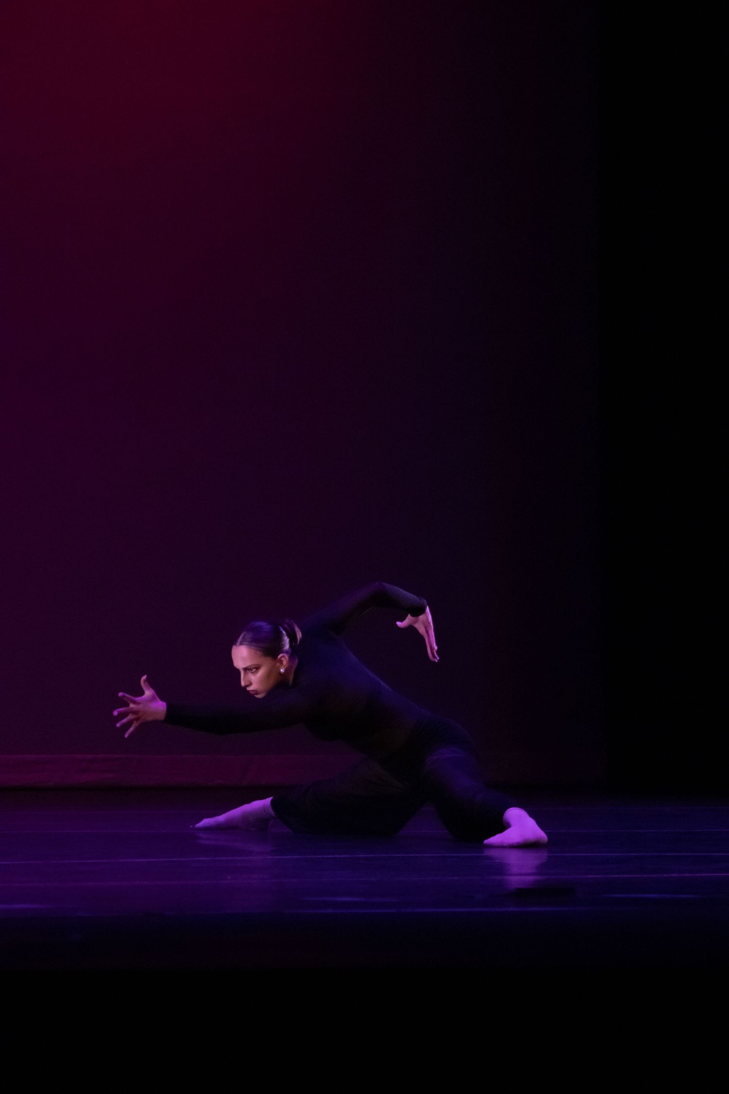 A dancer in a black bodysuit performs a low dramatic floor pose on a dark purple-lit stage.