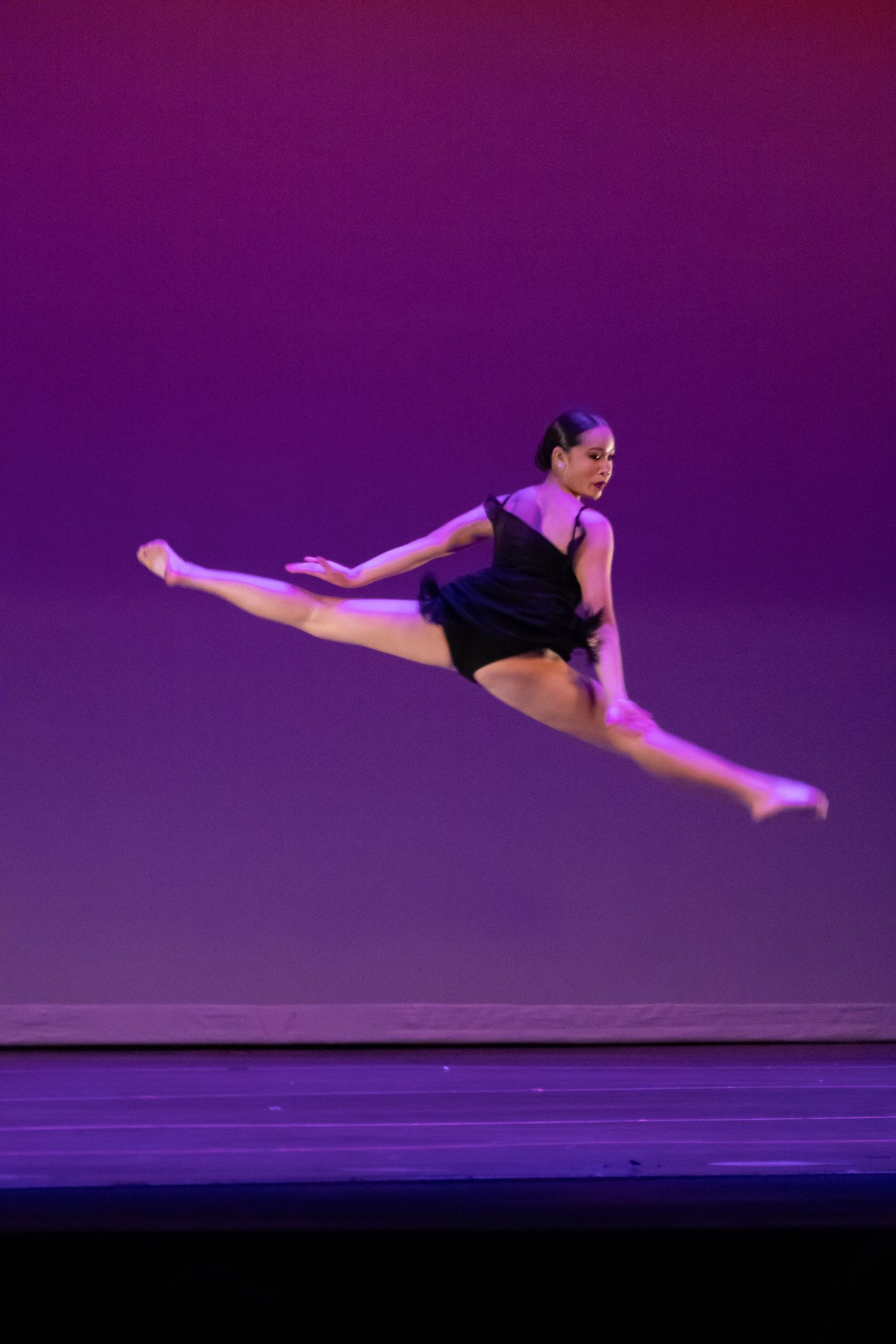 A dancer leaps gracefully into a split jump mid-air against a purple-pink stage backdrop.