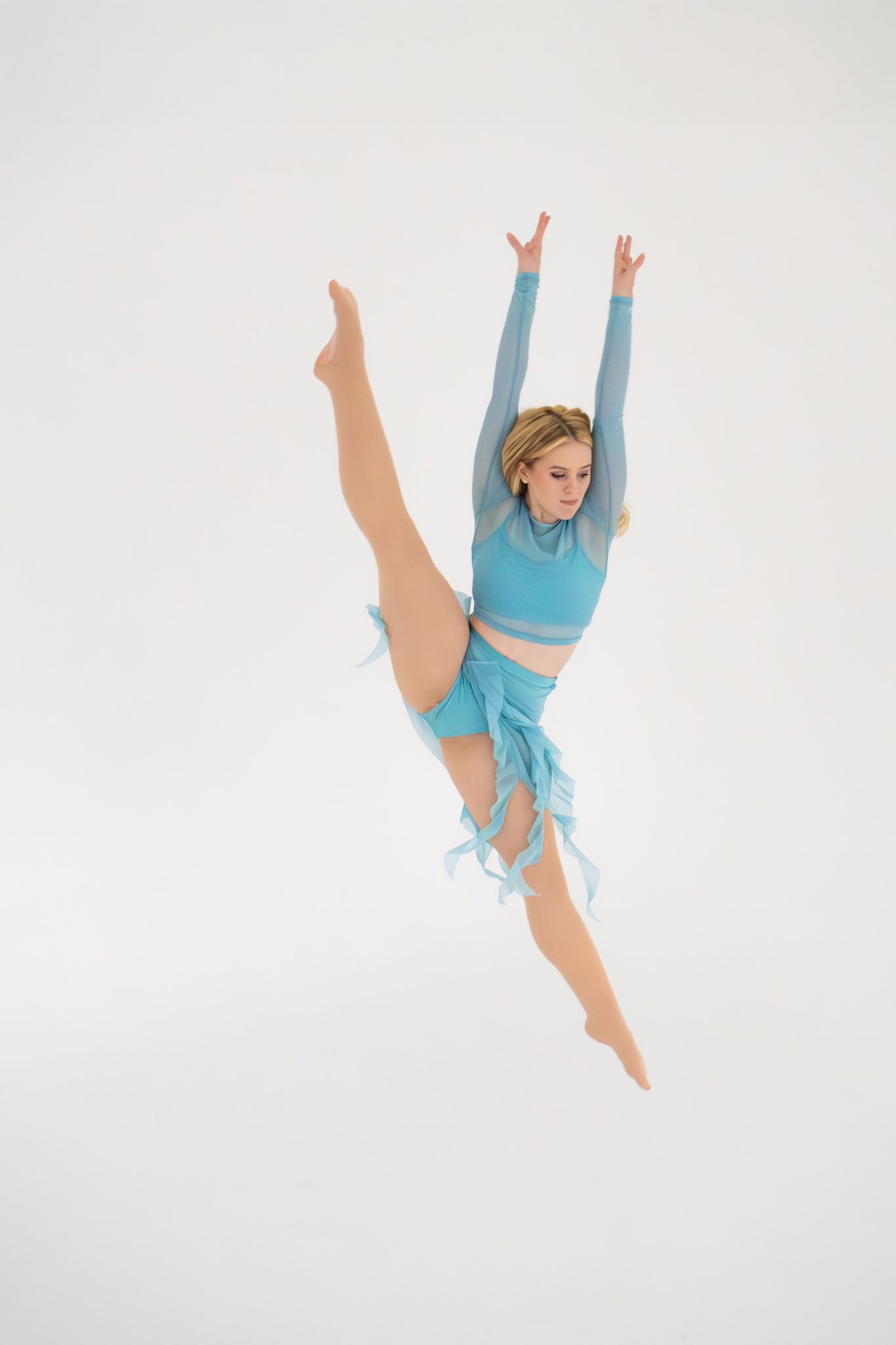 Blonde dancer in turquoise outfit performing aerial split jump against white background.