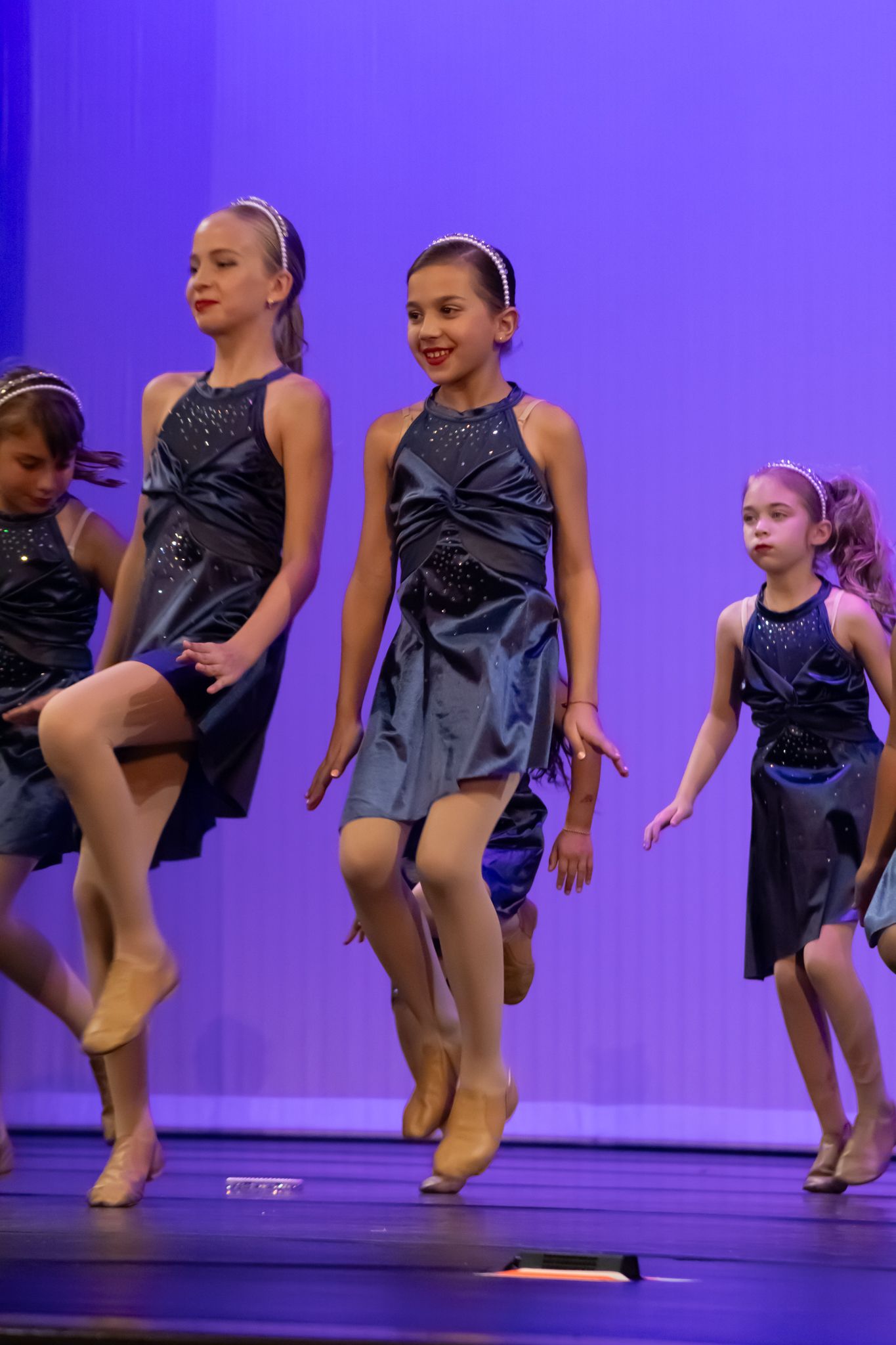 Young girls in sparkly navy blue dresses perform an energetic jazz dance routine on a blue-lit stage.