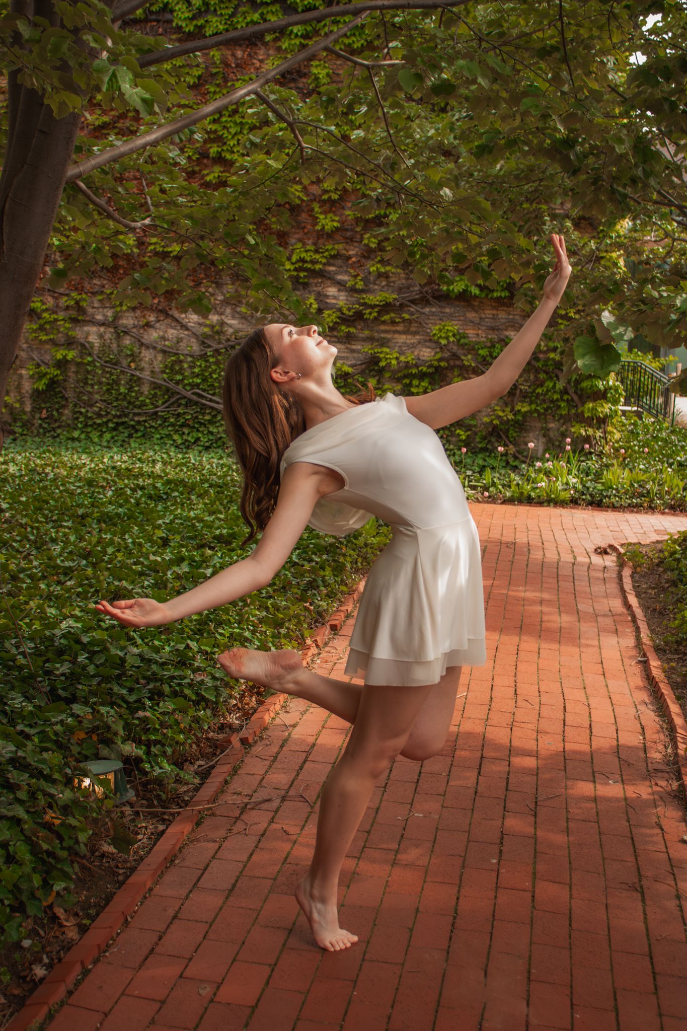 Barefoot dancer in white dress performing graceful ballet pose on brick path surrounded by lush green garden.