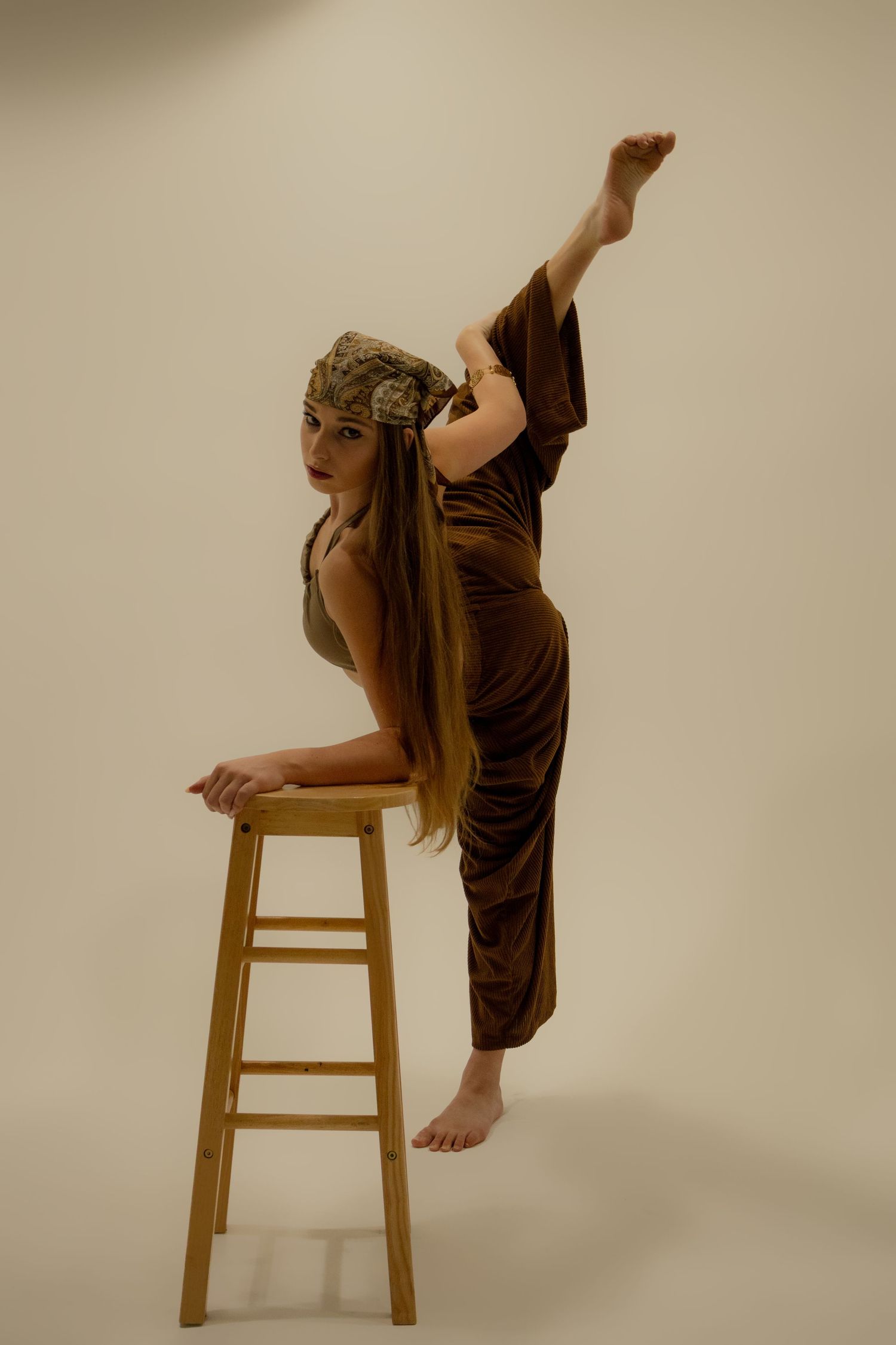 Flexible dancer in brown velvet outfit performs high leg extension beside wooden stool in studio.