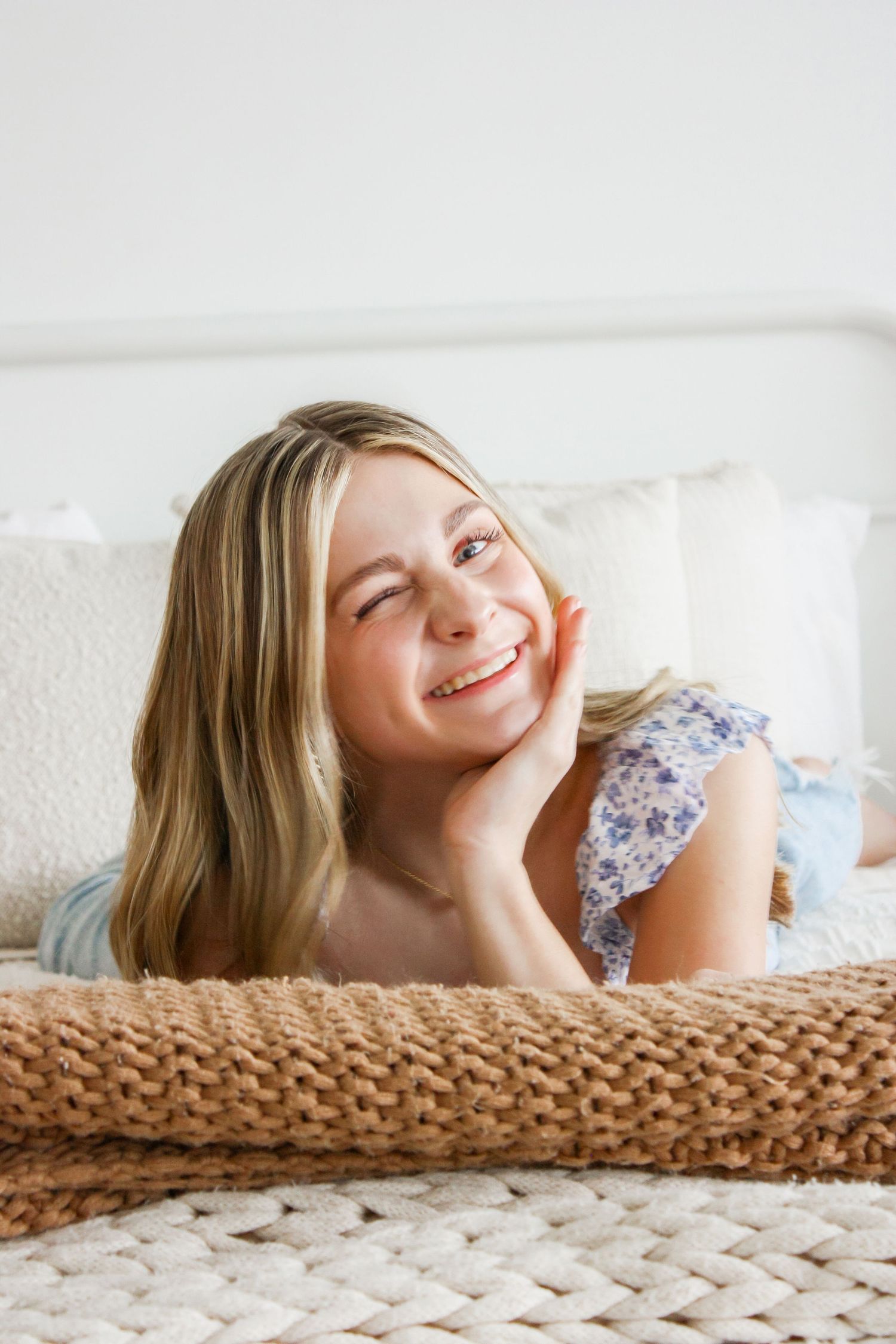 Smiling blonde woman lying on a bed with woven blanket, resting her chin on her hand in a bright room.