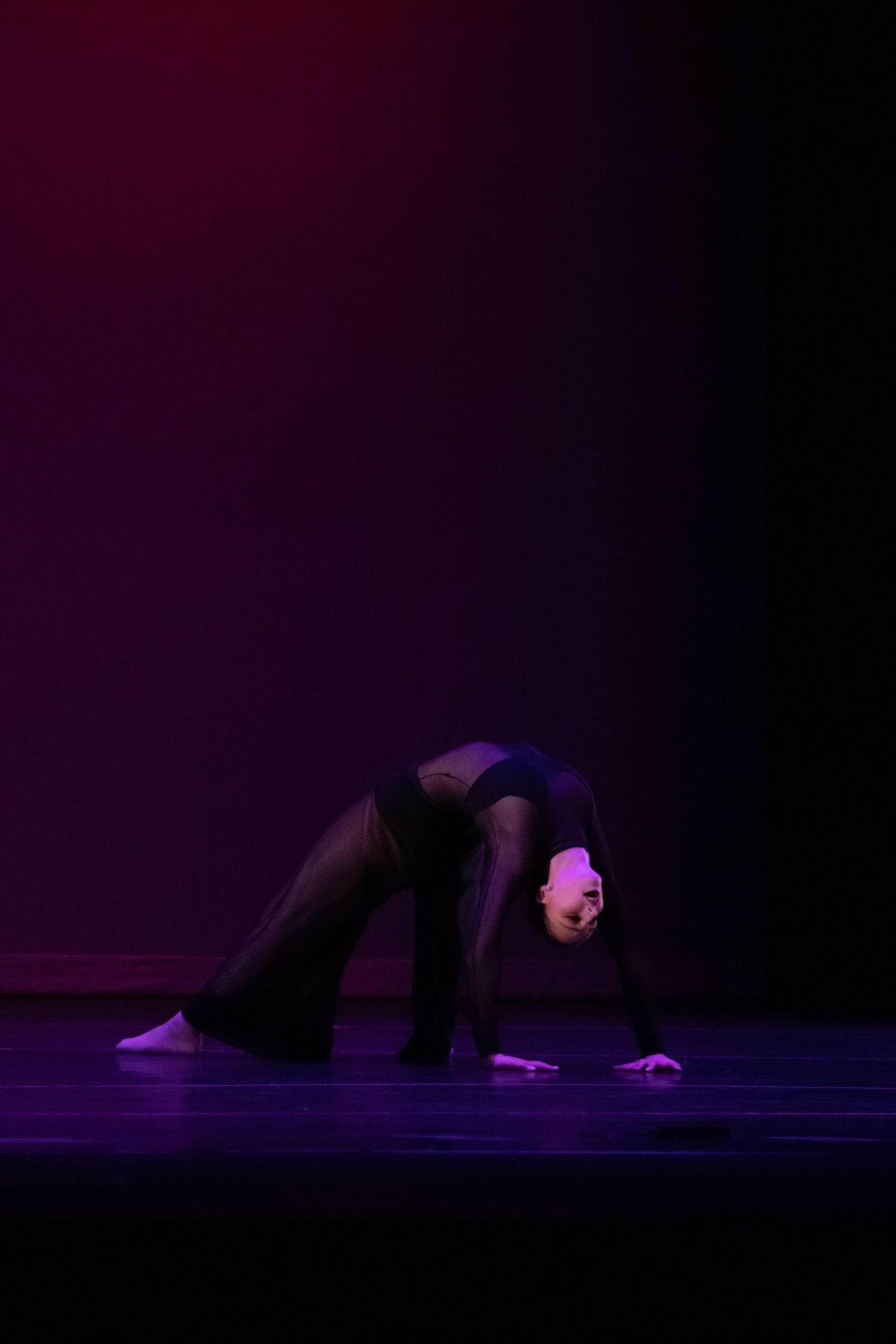 A dancer in black performs a dramatic backbend on a dimly lit stage with purple lighting.