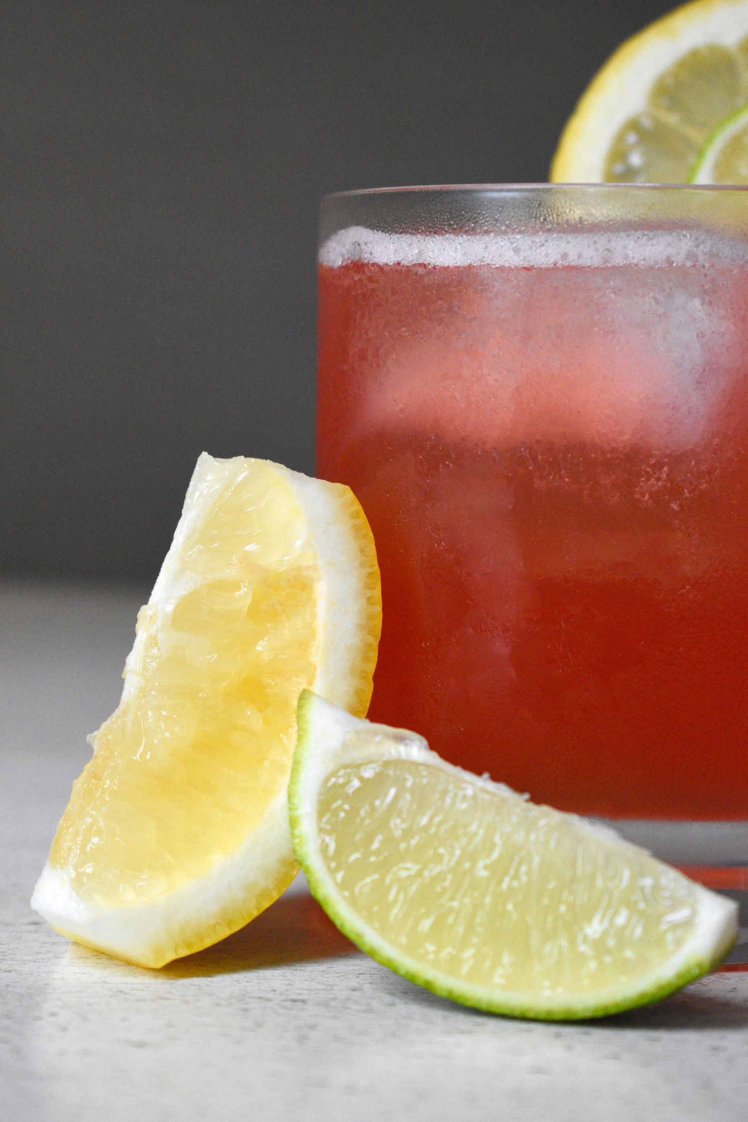 A refreshing red cranberry cocktail with ice, garnished with lemon and lime wedges on a gray surface.