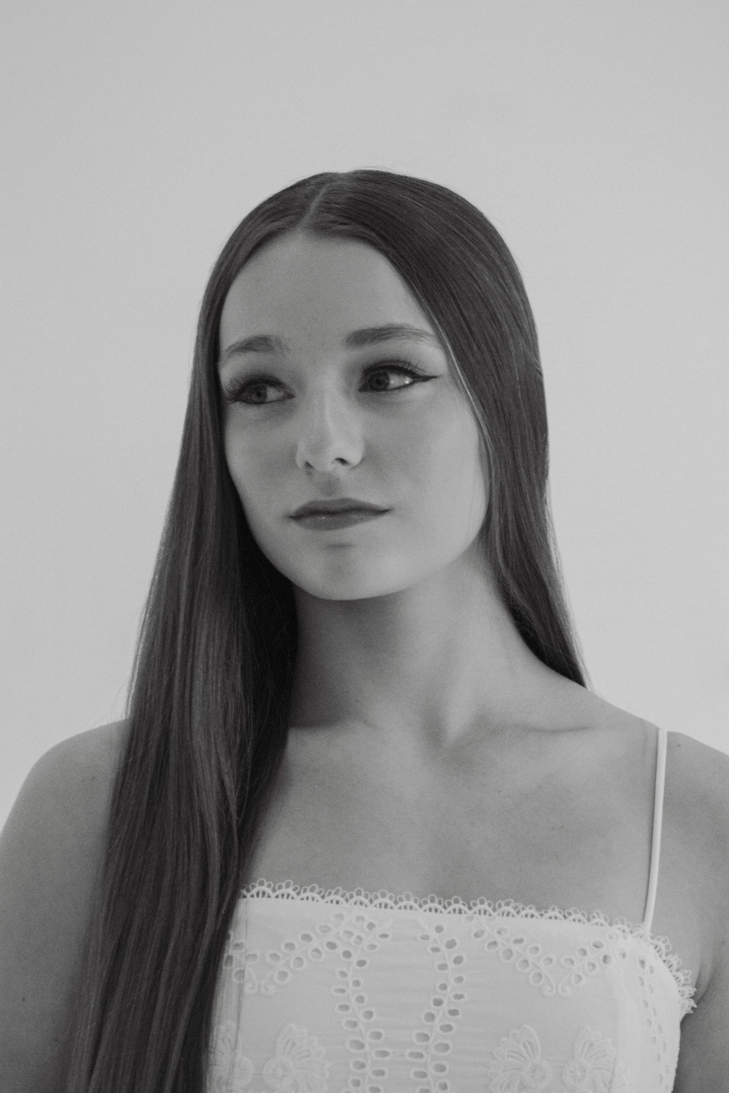 Young woman with long straight dark hair wearing a white lace dress in a black and white studio portrait.