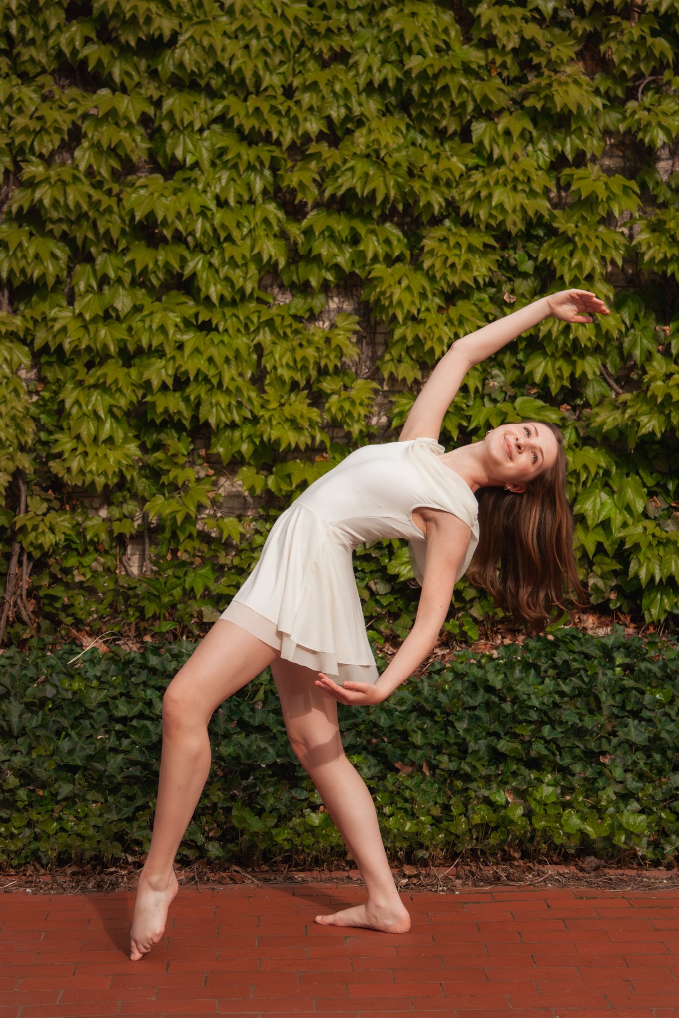 A young dancer in a white dress performs a side bend stretch against a green ivy-covered wall outdoors.