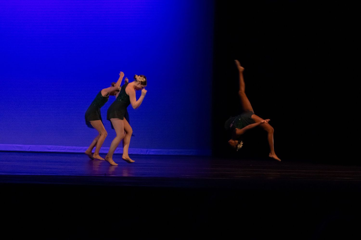 Three contemporary dancers perform on a dimly lit stage with blue and purple lighting during a recital.
