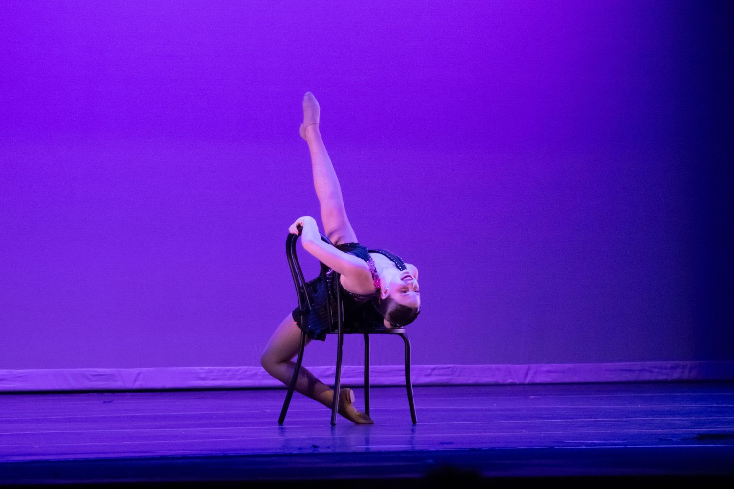 A flexible dancer performs an impressive backbend over a chair with legs raised under purple stage lights.