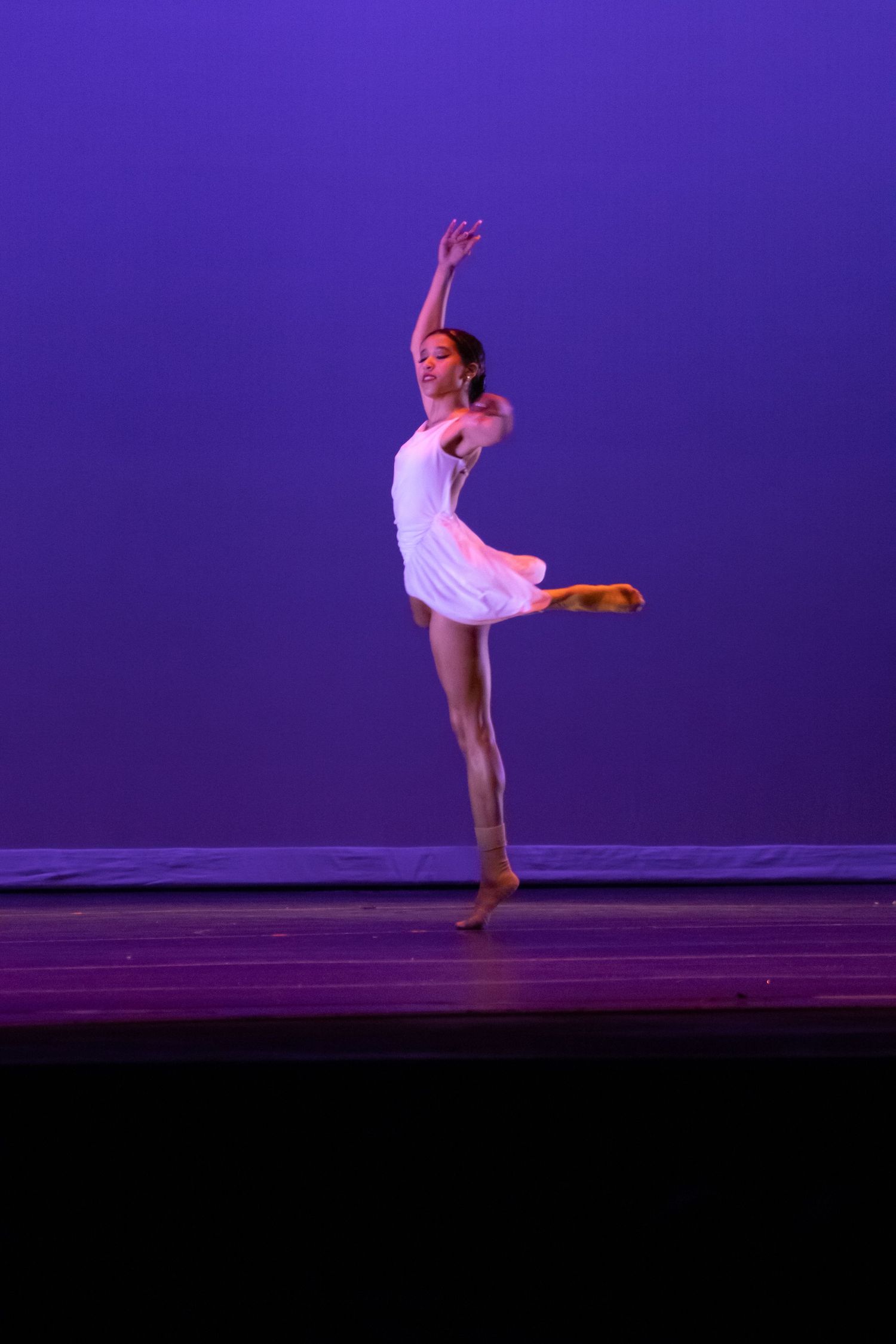 A young ballet dancer in a white leotard performs an elegant arabesque under purple stage lighting.