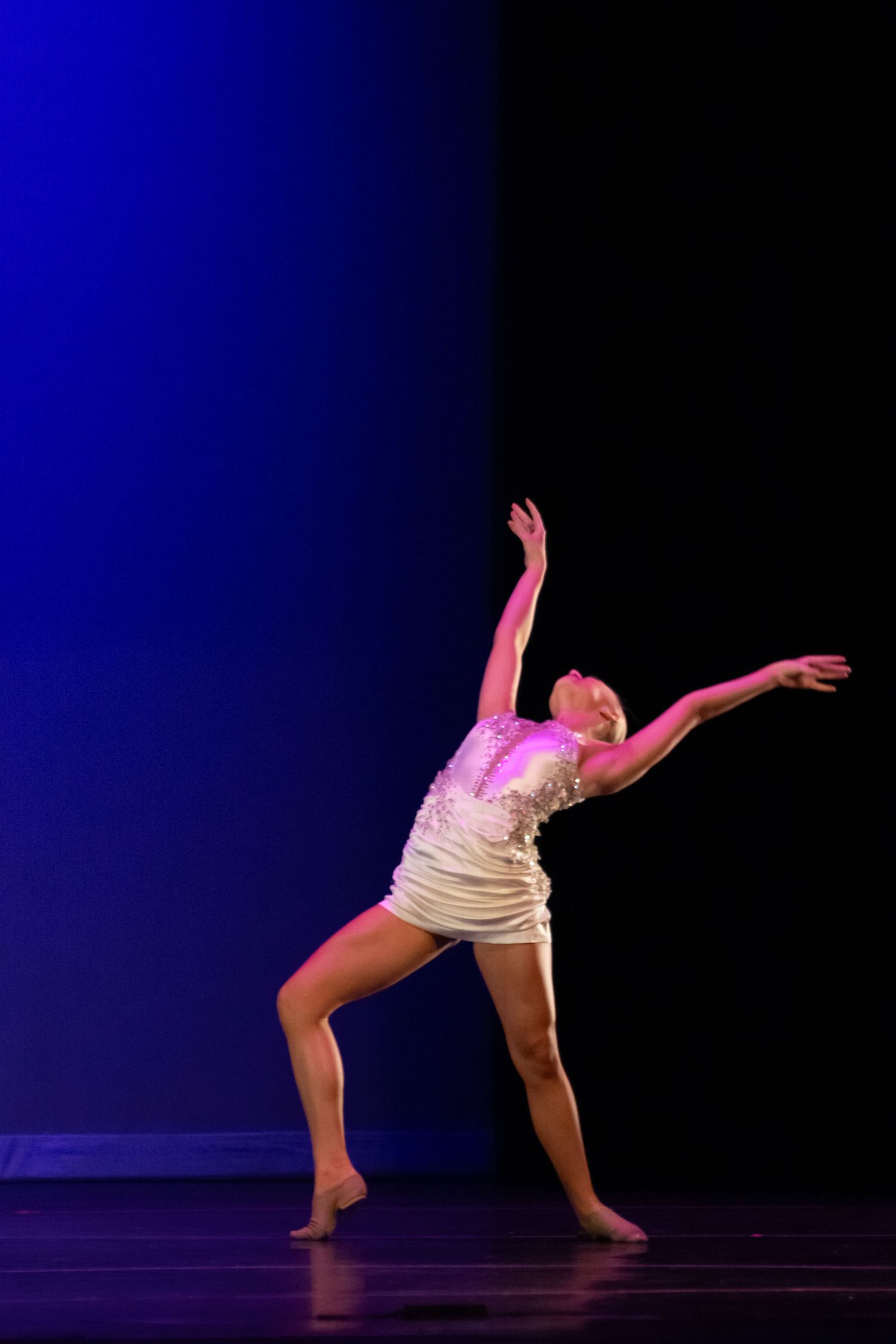 A female dancer in a white sequin dress strikes an expressive pose under pink and blue stage lights.