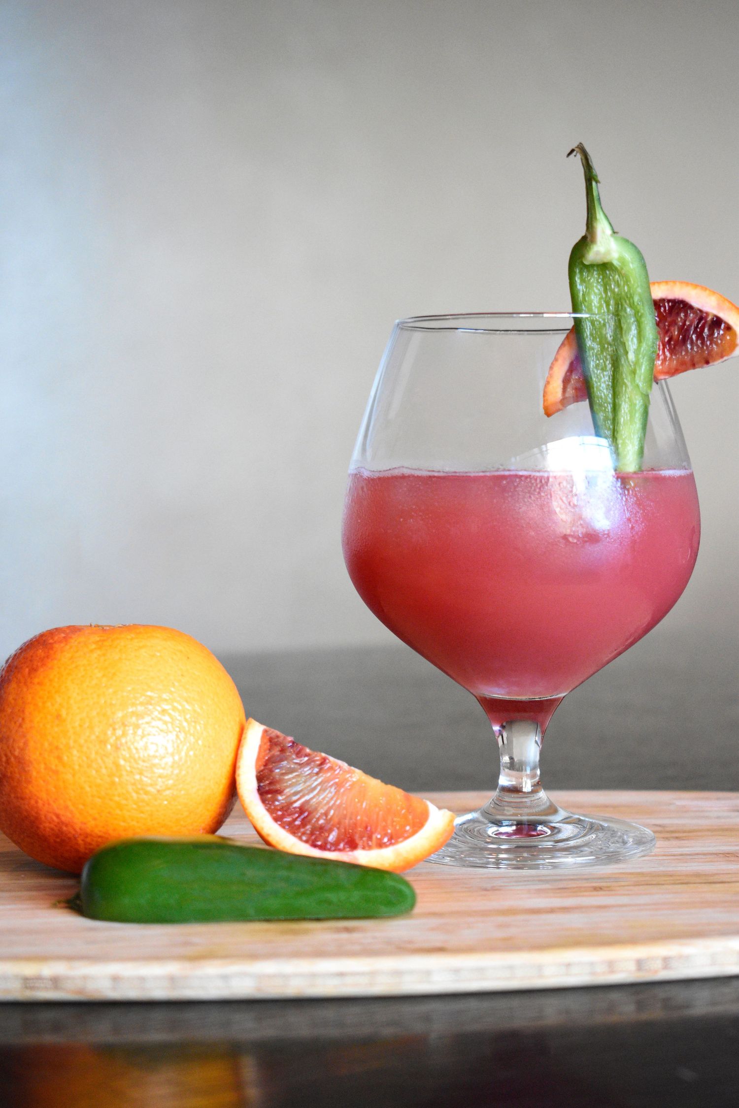 Blood orange cocktail in a wine glass garnished with a green pepper on a cutting board.