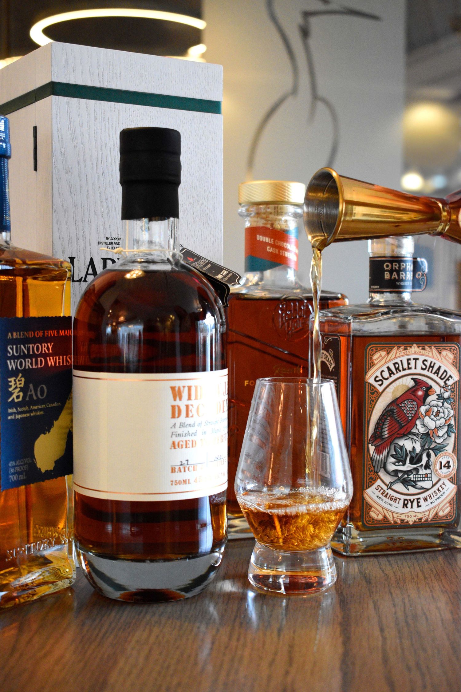 A bottle of Widow Jane Decadence whiskey being poured into a Glencairn glass surrounded by premium spirit bottles.