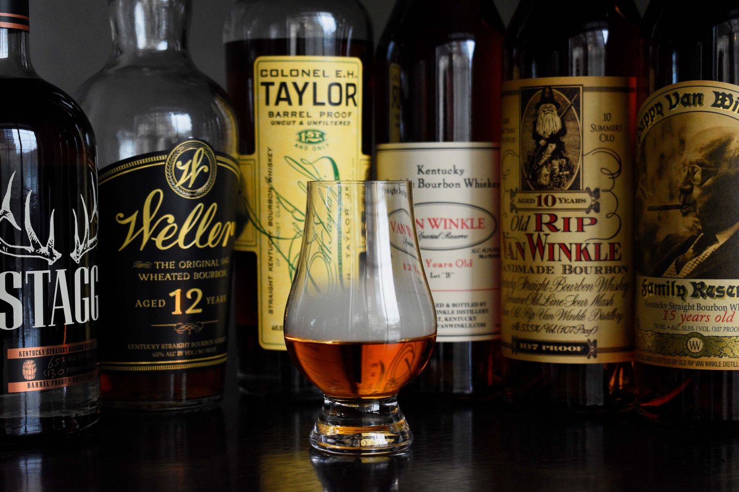 Glencairn glass of bourbon with Colonel Taylor, Weller, and Pappy Van Winkle bottles in background.