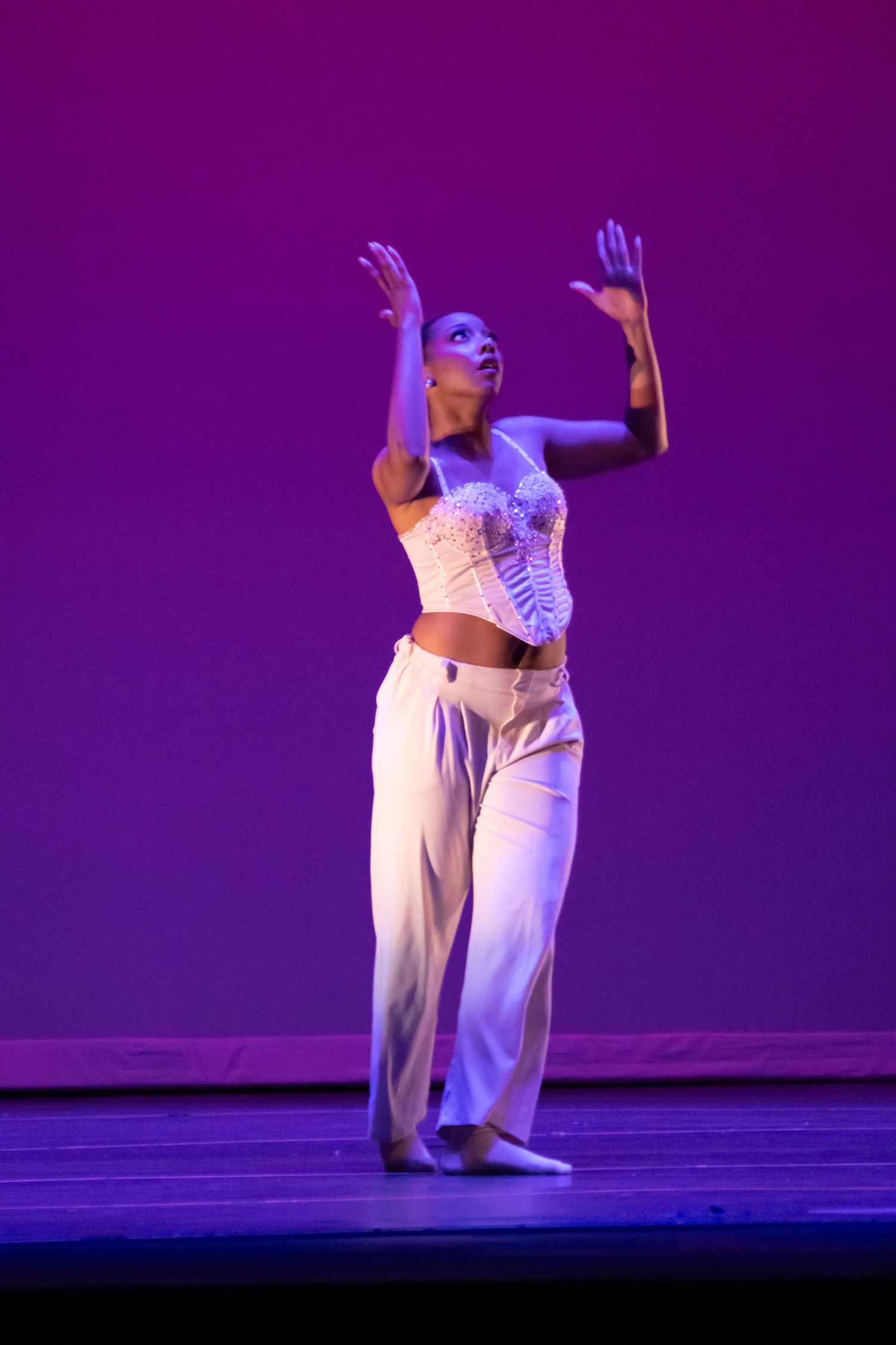A contemporary dancer in white raises both arms upward in an expressive pose on a purple-lit stage.