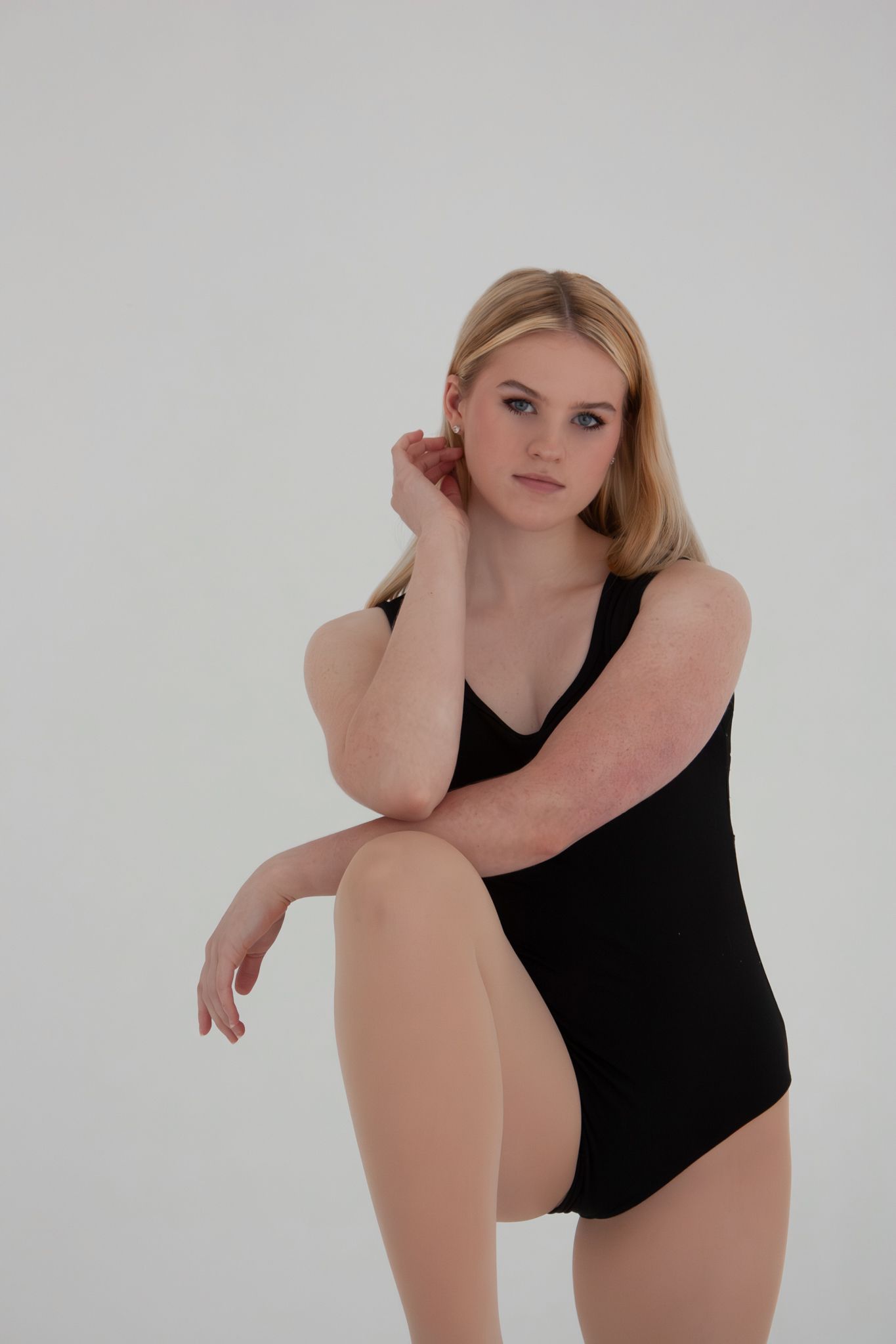Young blonde woman in black bodysuit posing seated against white background.